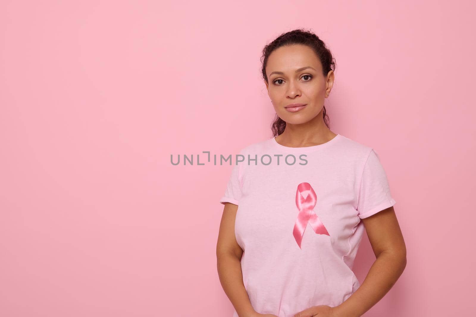 Confident portrait of a serene young mixed race African woman wearing a pink t-shirt with pink satin ribbon in symbol of International Day of Cancer Awareness. Oncological diseases concept. Copy space by artgf