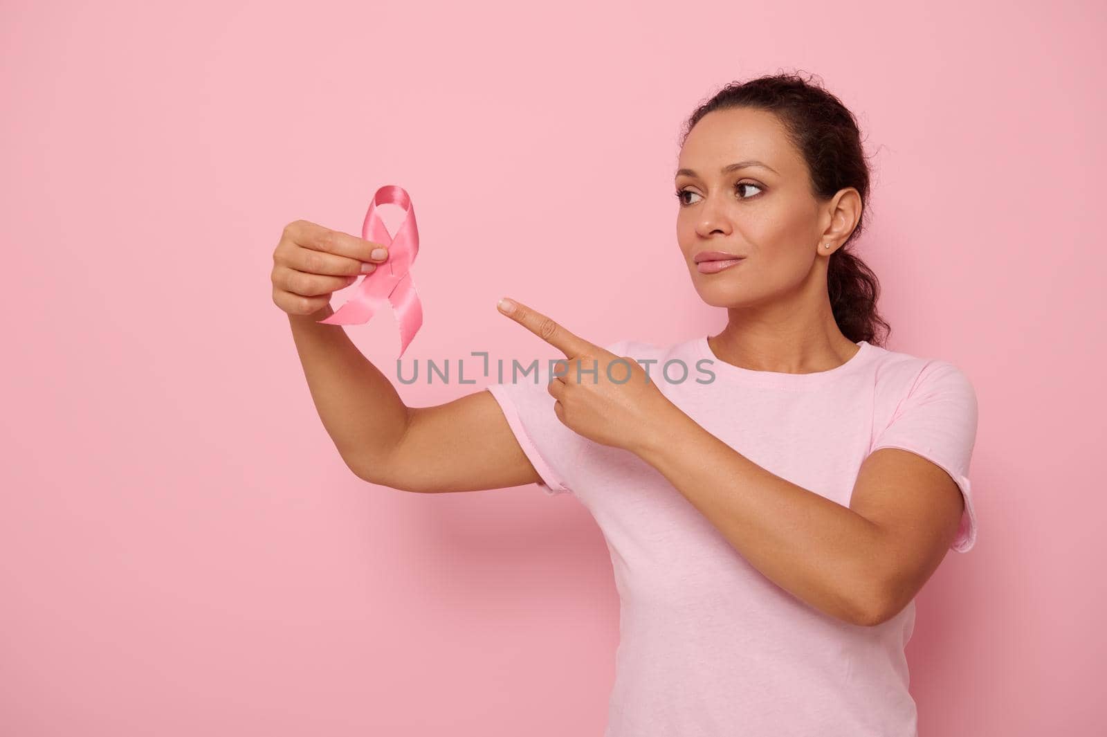 Beautiful mixed race woman wearing pink T-shirt pointing to a pink satin ribbon in her hand, colored background copy space. Concept of 1 st October, International Day of fight Breast Cancer disease by artgf