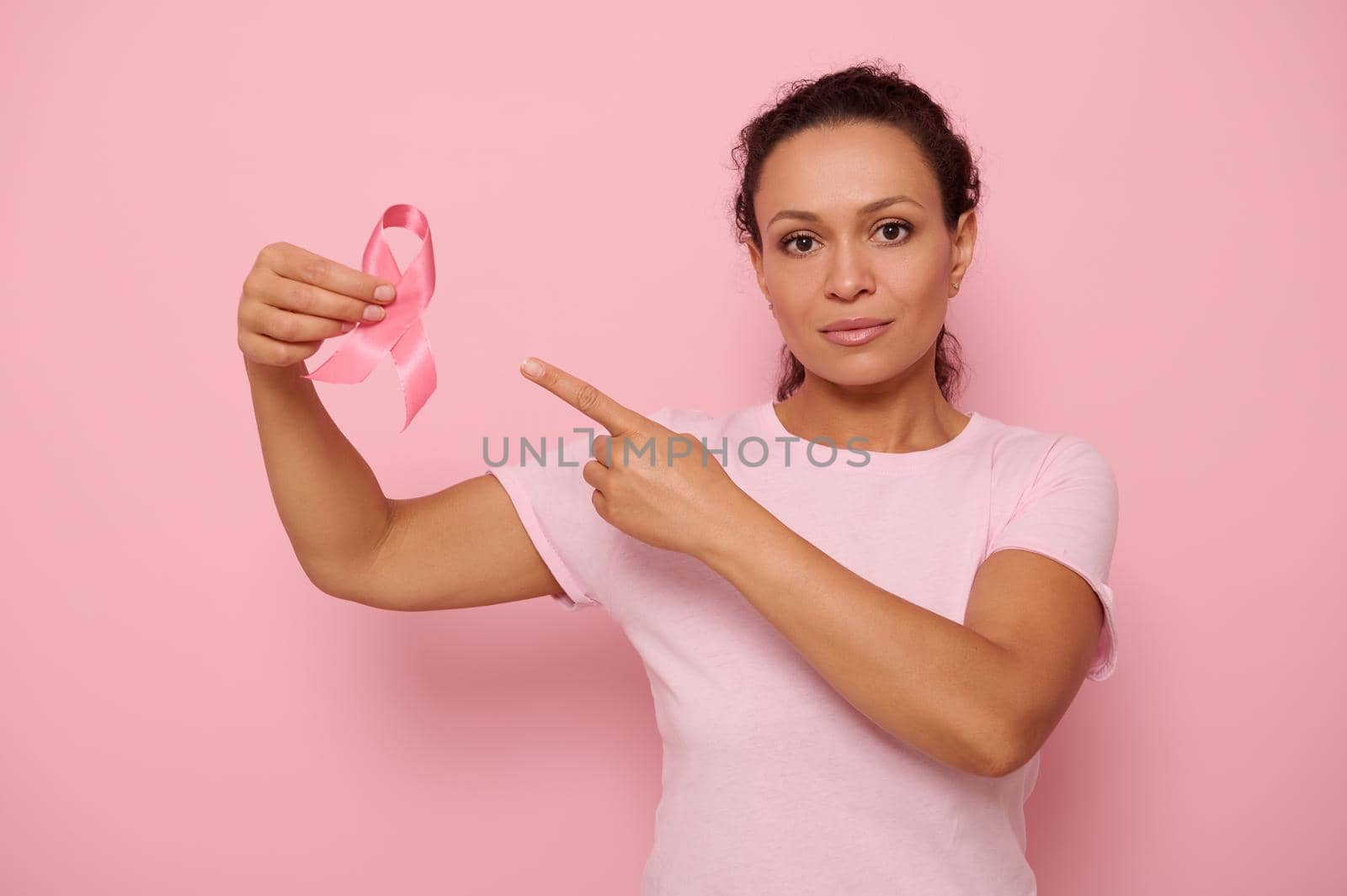 Portrait of serene Middle Eastern ethnicity woman in pink T-shirt pointing to a satin ribbon in her hand, looking at camera, isolated on colored background with copy space. Breast Cancer Day concept by artgf