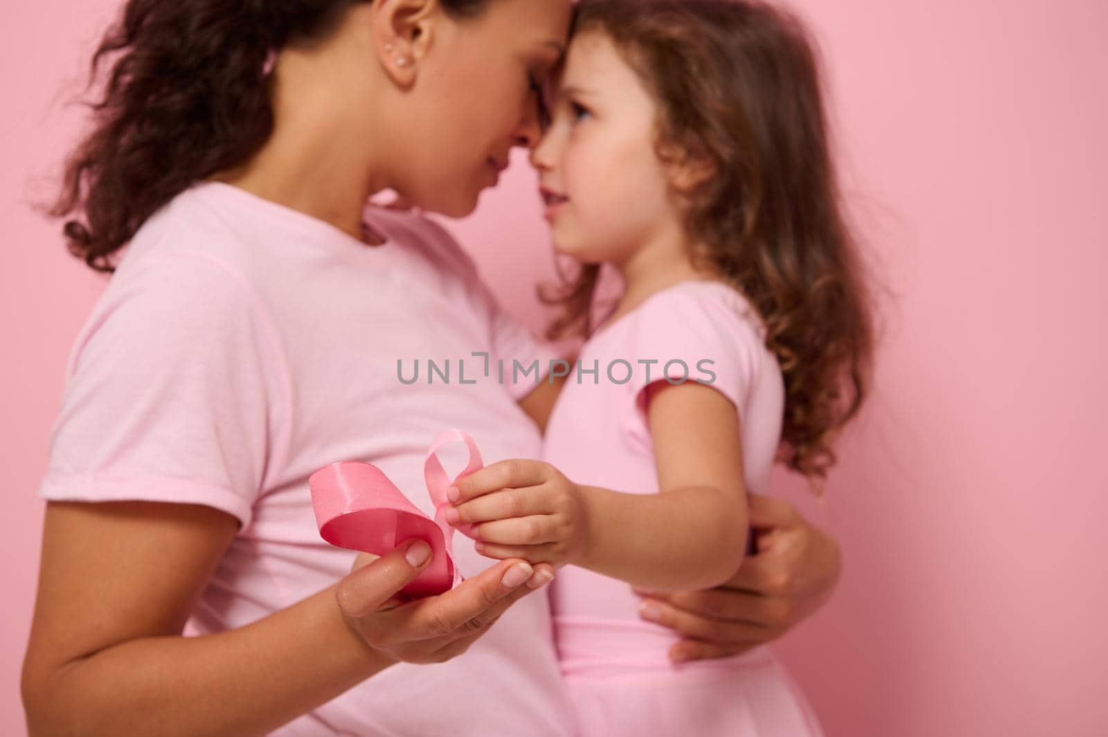 Focus on pink satin ribbon, symbol of International Breast Cancer Awareness Day, against blurred background of loving mother and lovely daughter. Female healthcare and medical education concept