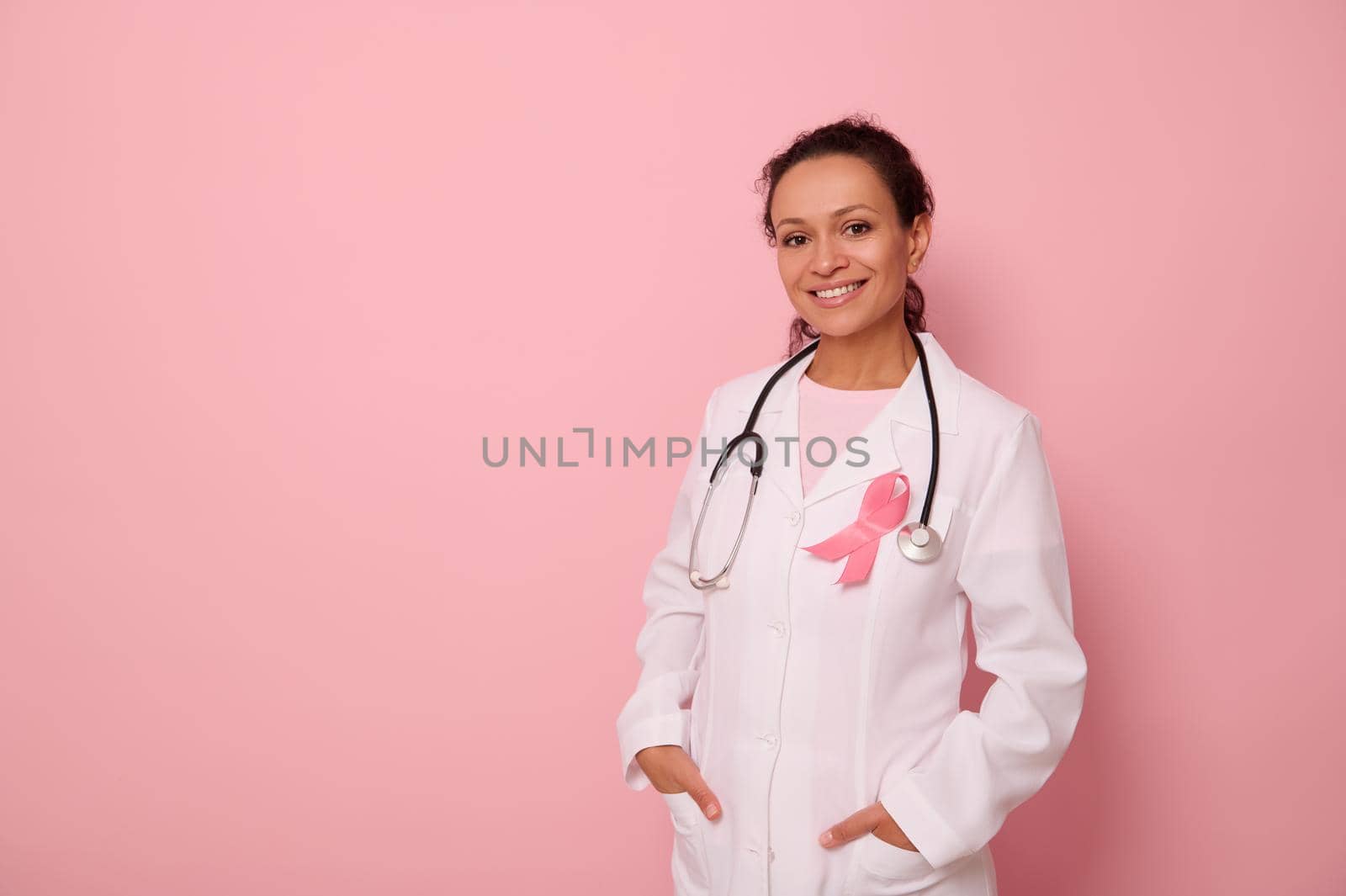 Portrait of African American womandoctor in medical coat and pink ribbon, stethoscope around neck, looking at camera, isolated on colored background, copy space. World Breast Cancer Awareness Day