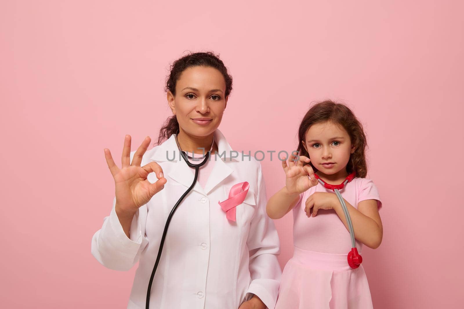 Pretty woman doctor in medical gown and a cute little girl, both with pink ribbon and phonendoscope around their neck, gesticulate with an OK sign to support breast cancer patients and survivors. by artgf