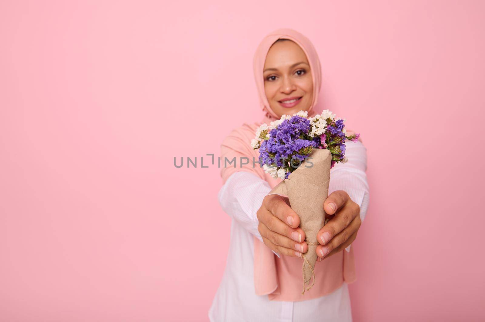 Focus on a beautiful simple bouquet of meadow wildflowers in purple tones, wrapped in brown craft paper in the hands of an attractive smiling Muslim woman wearing a hijab. Pink background copy space