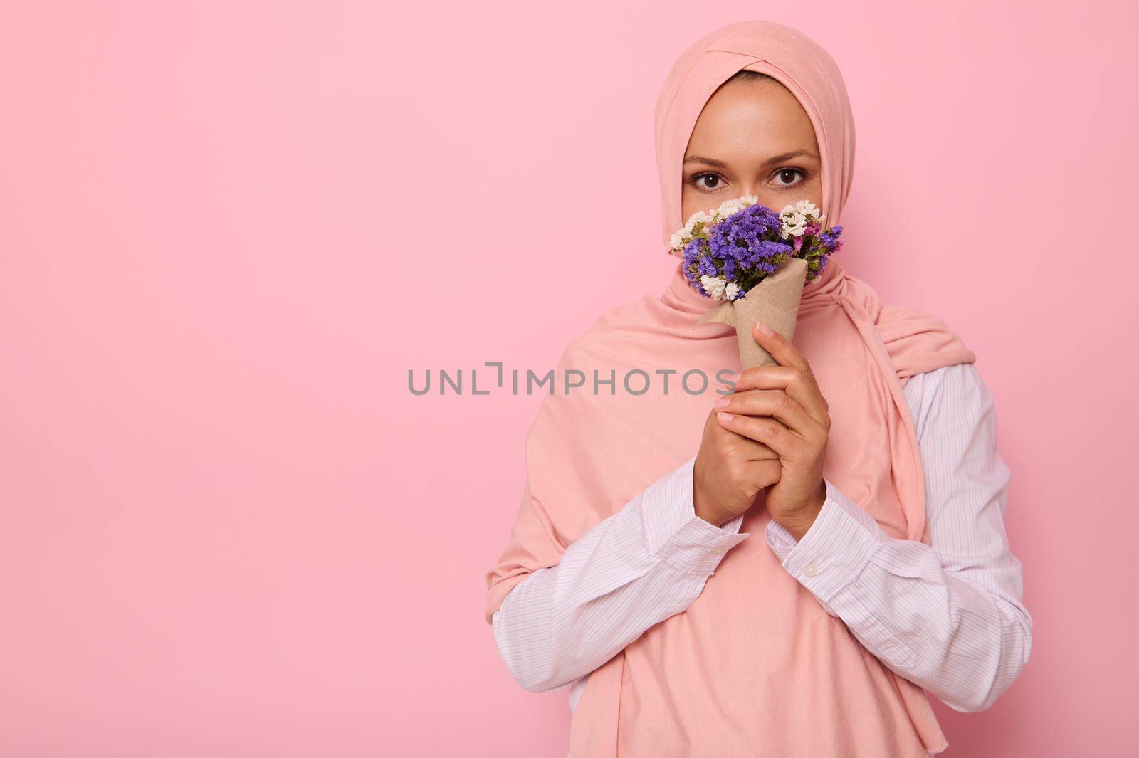 Confident portrait of young charming Arab Muslim woman in pink hijab with beautiful dark eyes, attractive gaze, looking at camera, covers half of her face and mouth with a craft bouquet of wildflowers