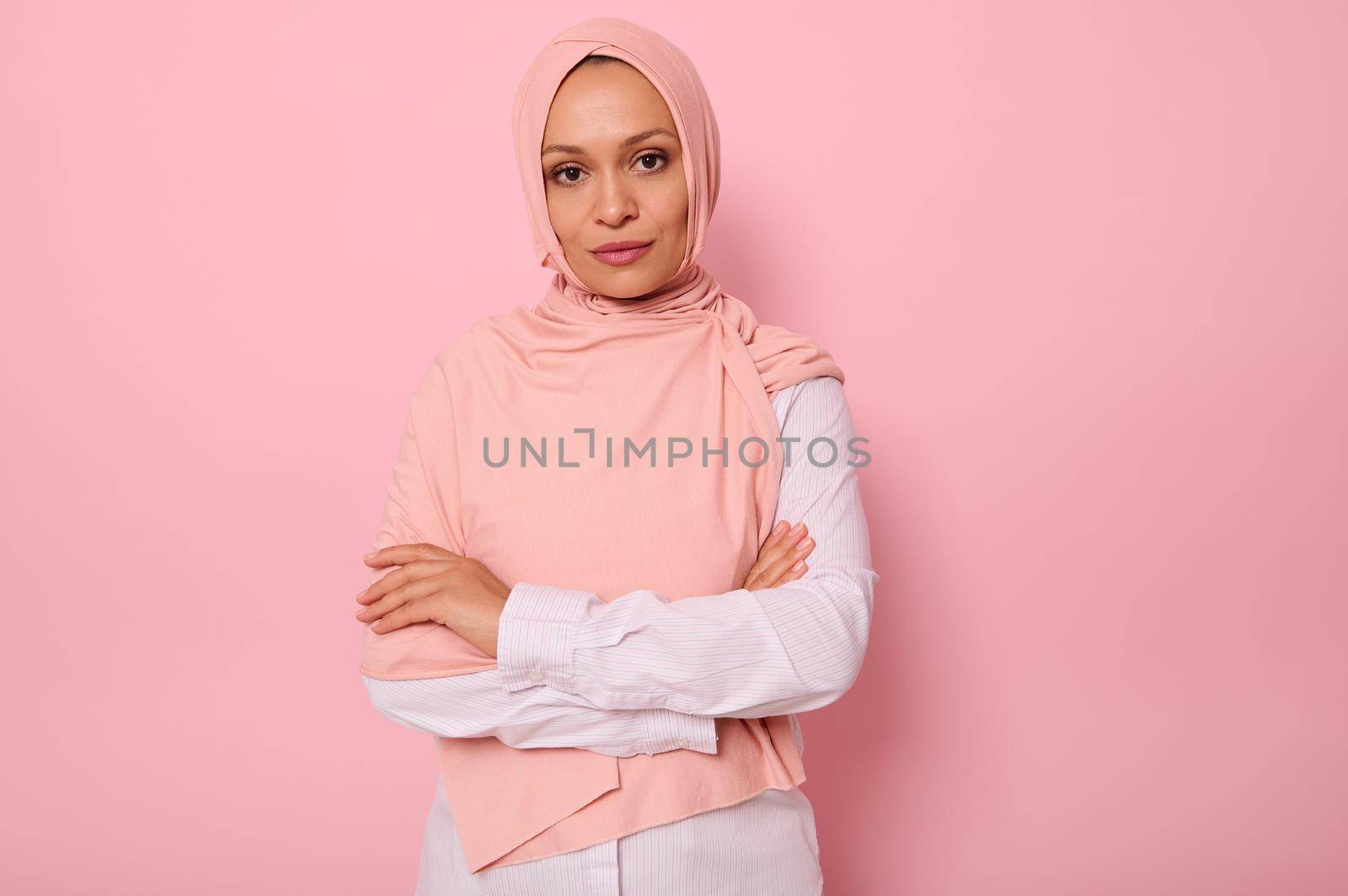 Waist-length portrait of beautiful Arabic Muslim woman in pink hijab posing looking to camera with attractive gaze, confident look and crossed arms, on pink background with copy space