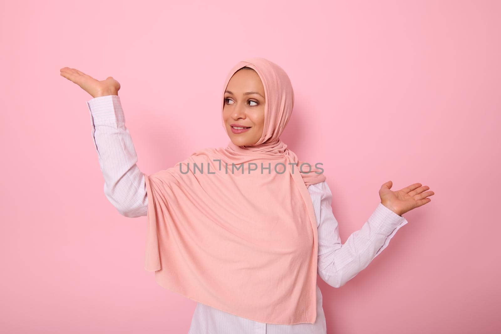 Beautiful young Arab Muslim woman raising hand palm up while looking at it with smile. Isolated background with copy space