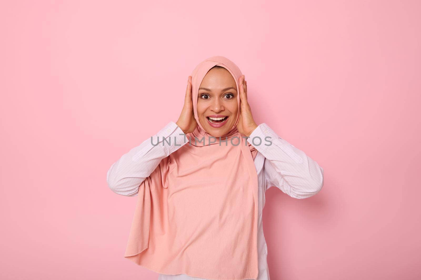 Main focus on facial expression of Muslim woman in pink hijab holding her head with hands, smiling with toothy smile looking at camera with surprised look, isolated on colored background, copy space.