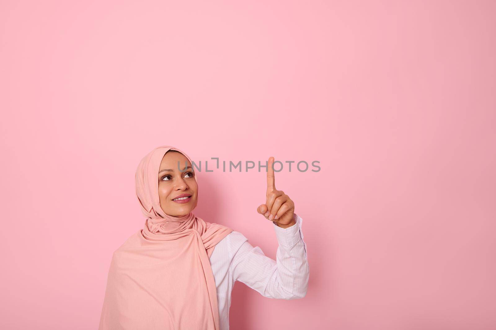 Beautiful Muslim woman of Middle Eastern ethnicity dressed in religious outfit and covered head with hijab ethnicity smiles and looks up pointing her index finger up on a copy space of pink background