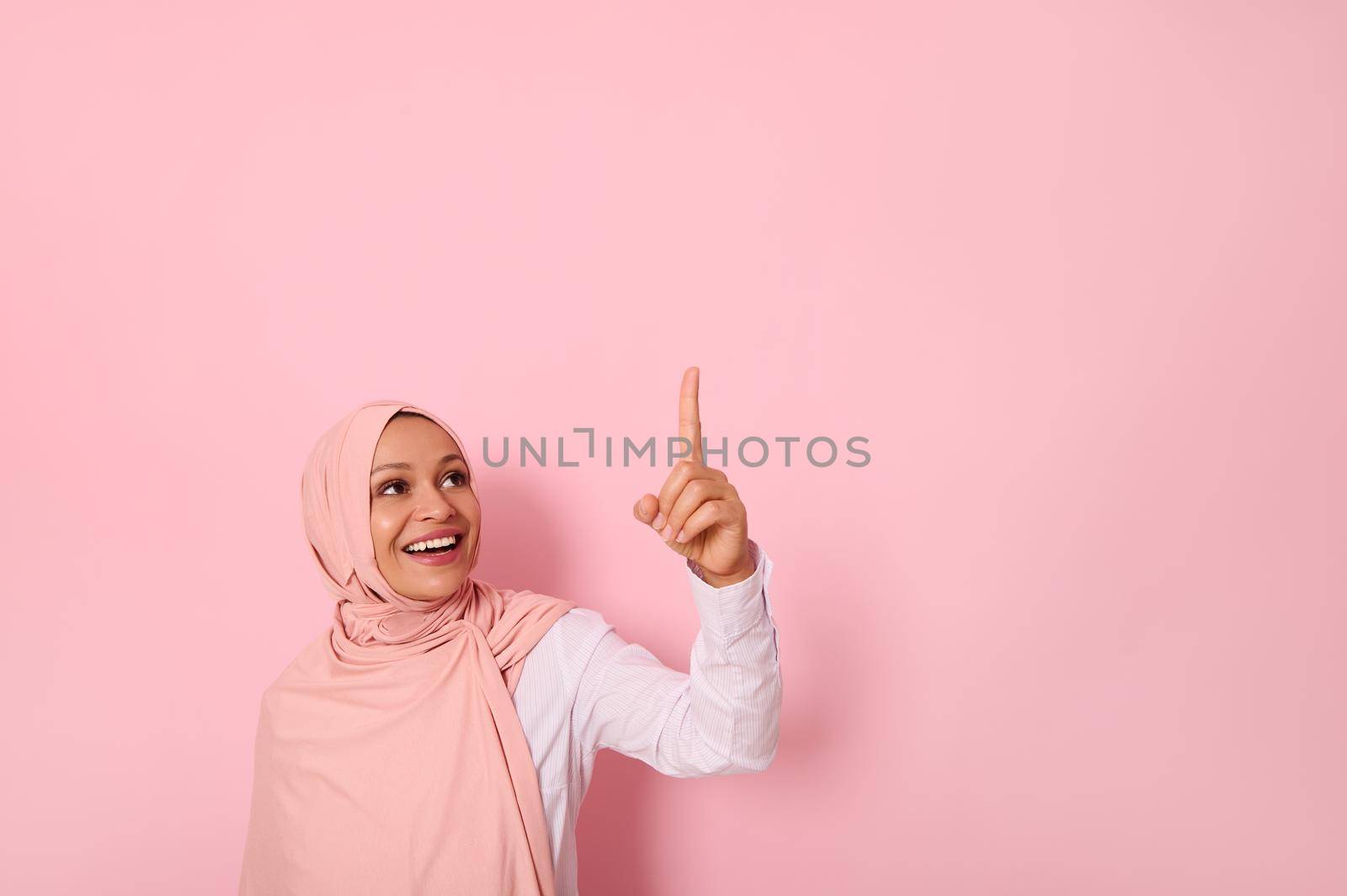 Muslim woman of Middle Eastern ethnicity dressed in religious outfit and covered head with hijab ethnicity smiles with toothy smile and looks up pointing her index finger on pink background copy space