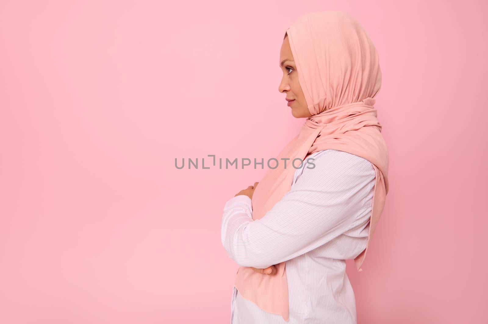 Confident close-up portrait of Arab muslim confident woman with attractive gaze , wearing a pink hijab looking confidently in front of herself standing sideways on colored background with copy space