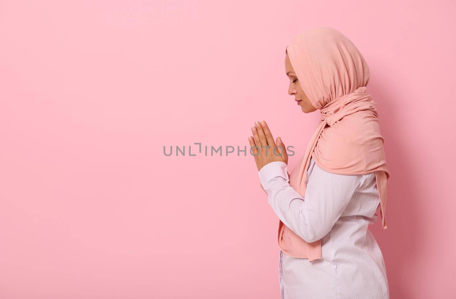 Side portrait of a serene Muslim Arab woman in pink hijab and strict outfit praying, performing namaz, isolated on colored background with space for text