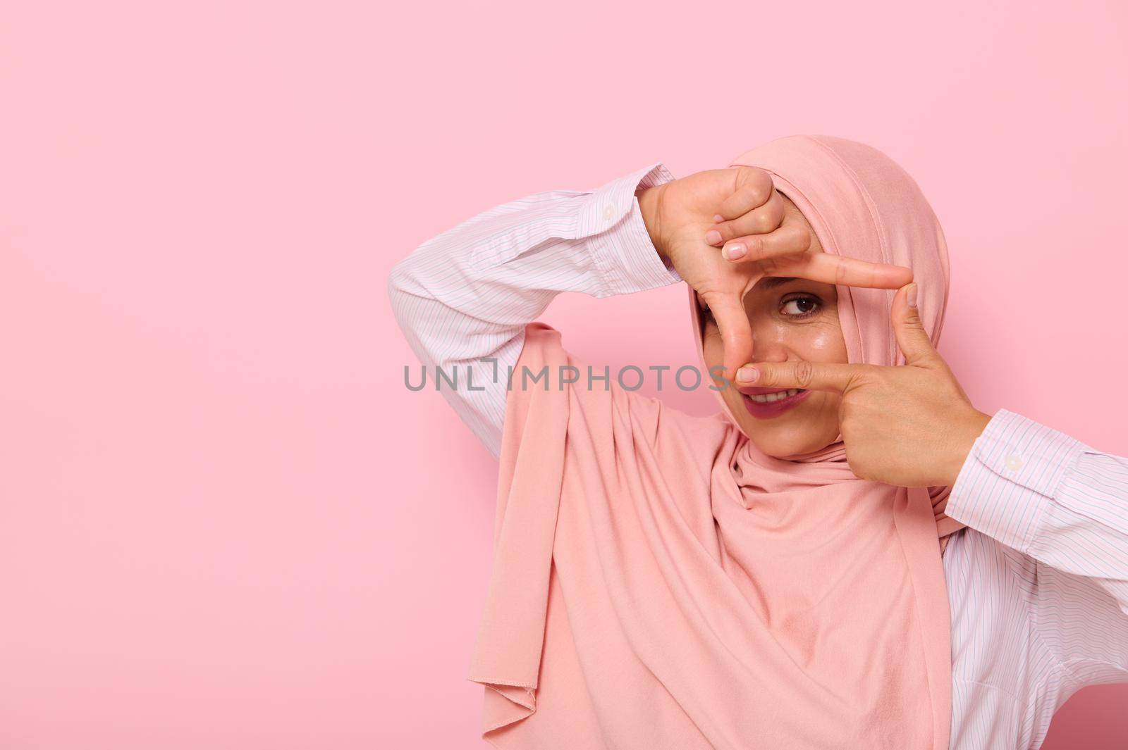 Headshot, close-up face portrait of a young Muslim pretty woman of Arabic ethnicity, wearing hijab and looking at the camera through a finger frame, isolated on colored background with copy space