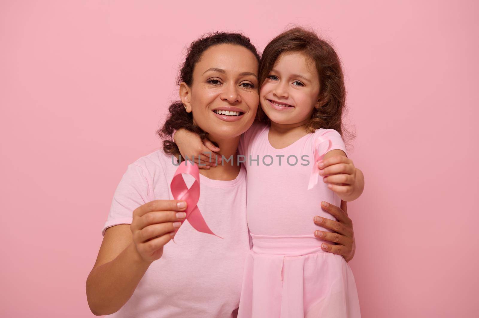 African American woman hugs her daughter, holding pink ribbon, cute smiles looking at camera, isolated on colored background with copy space. International Day of fight against a Breast Cancer disease by artgf
