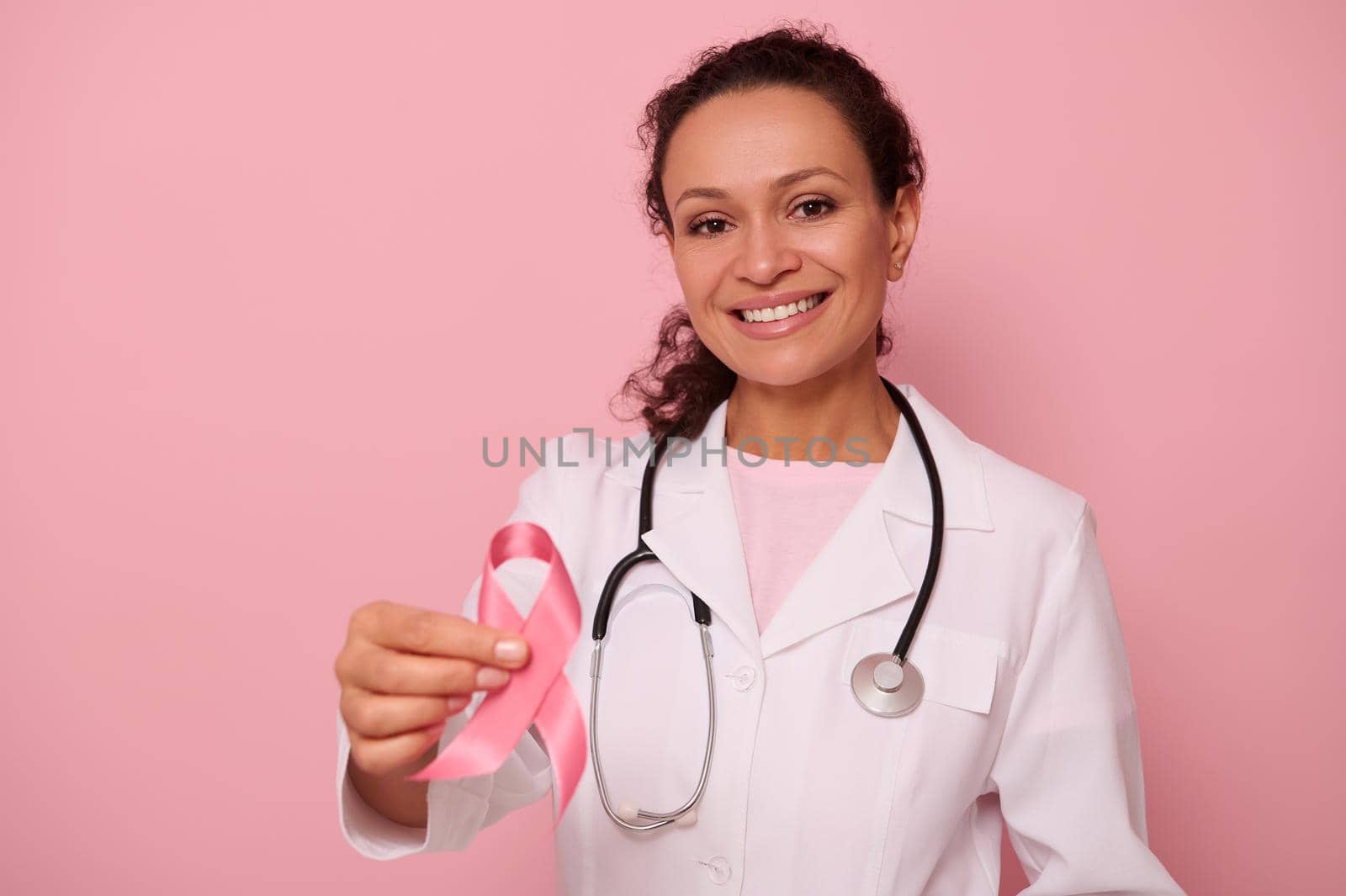 Gorgeous African American female doctor with beautiful toothy smile showing a Pink satin Ribbon, isolated on colored background with copy space. World Day of fight Breast Cancer, 1 st October concept by artgf