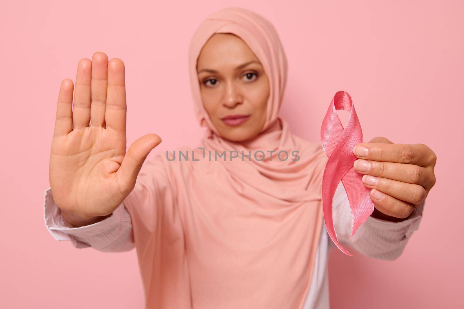 Confident portrait of blurred Muslim Arab woman in hijab holding a pink cancer awareness ribbon. Focus on woman's hand gesturing STOP , isolated over pink background with copy space. Fight Cancer Day