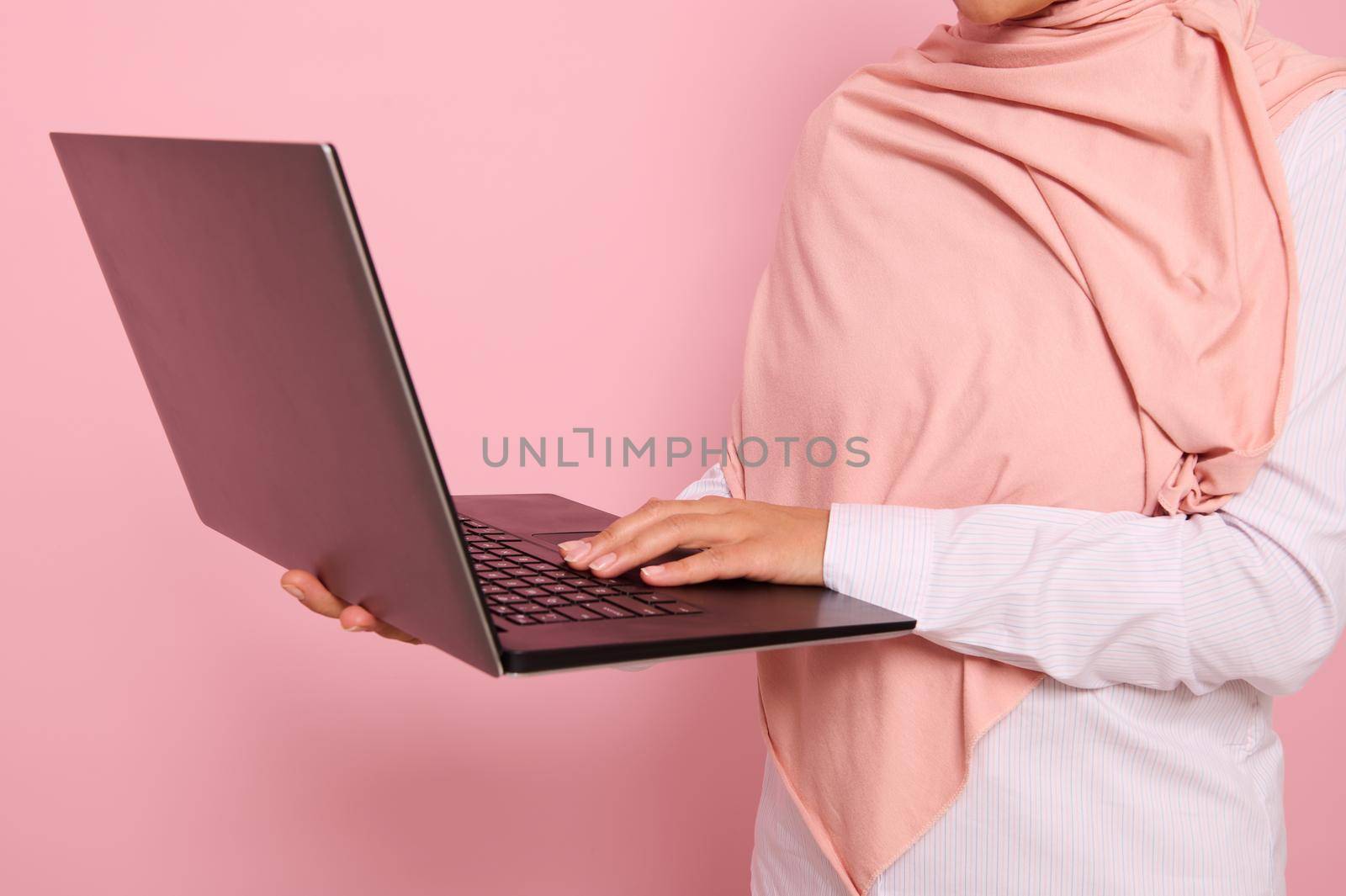 Close-up portrait of a woman wearing pink hijab standing against colored background with an open laptop in hands and typing text.Concept of successful Muslim programmer woman, worker at IT technology by artgf