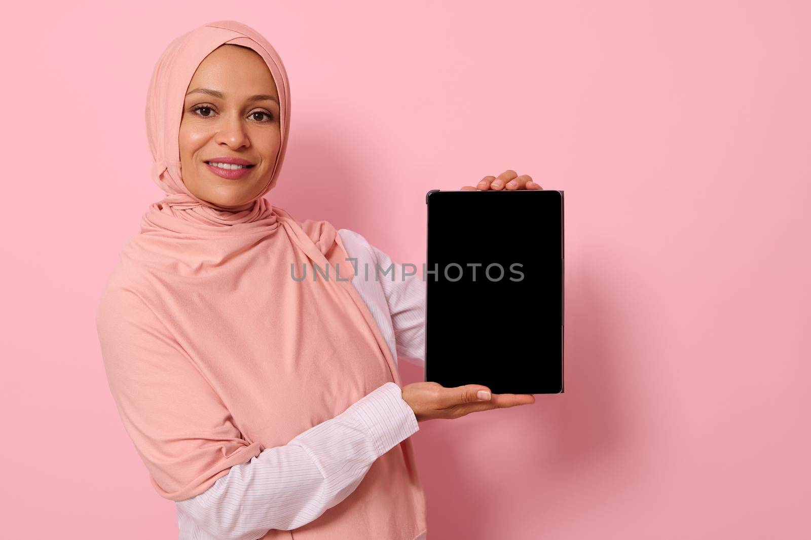 Confident Muslim woman of Arab or Middle Eastern ethnicity in hijab holding vertically a digital tablet and showing its blank screen, smiles looking at camera, isolated on pink background, copy space