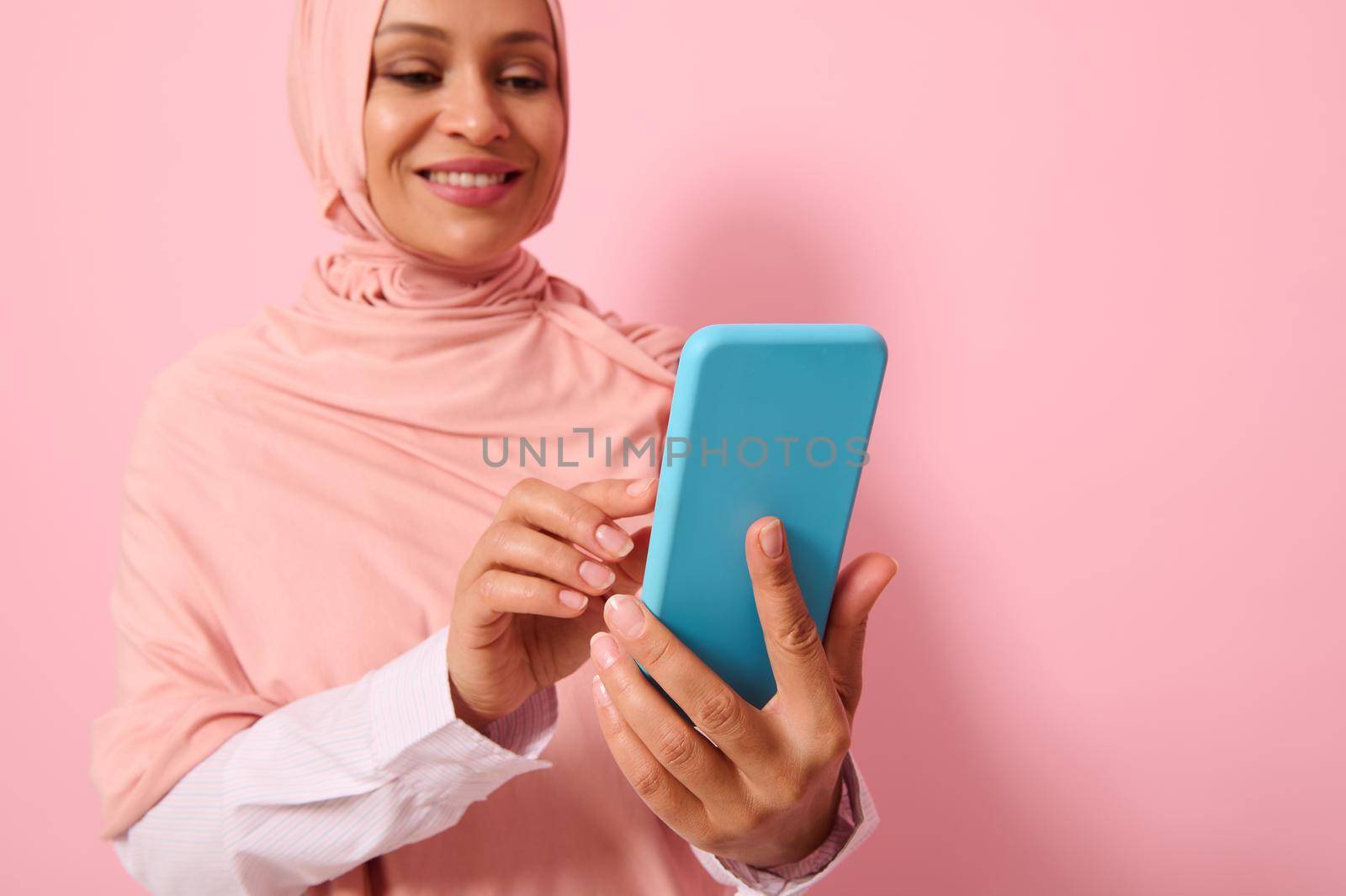Close-up portrait of confident Arab Muslim mature pretty woman in strict religious outfit and covered head in pink hijab texting message on a mobile phone in her hands, colored background, copy space