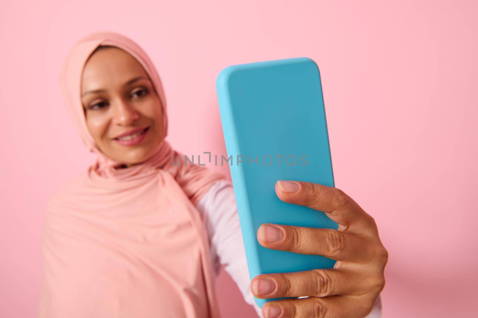 Soft focus on the smartphone in blue cover in outstretched arms of Arab Muslim woman wearing traditional religious islamic outfit, pink hijab,and smiles with toothy smile making a selfie, self-portrait
