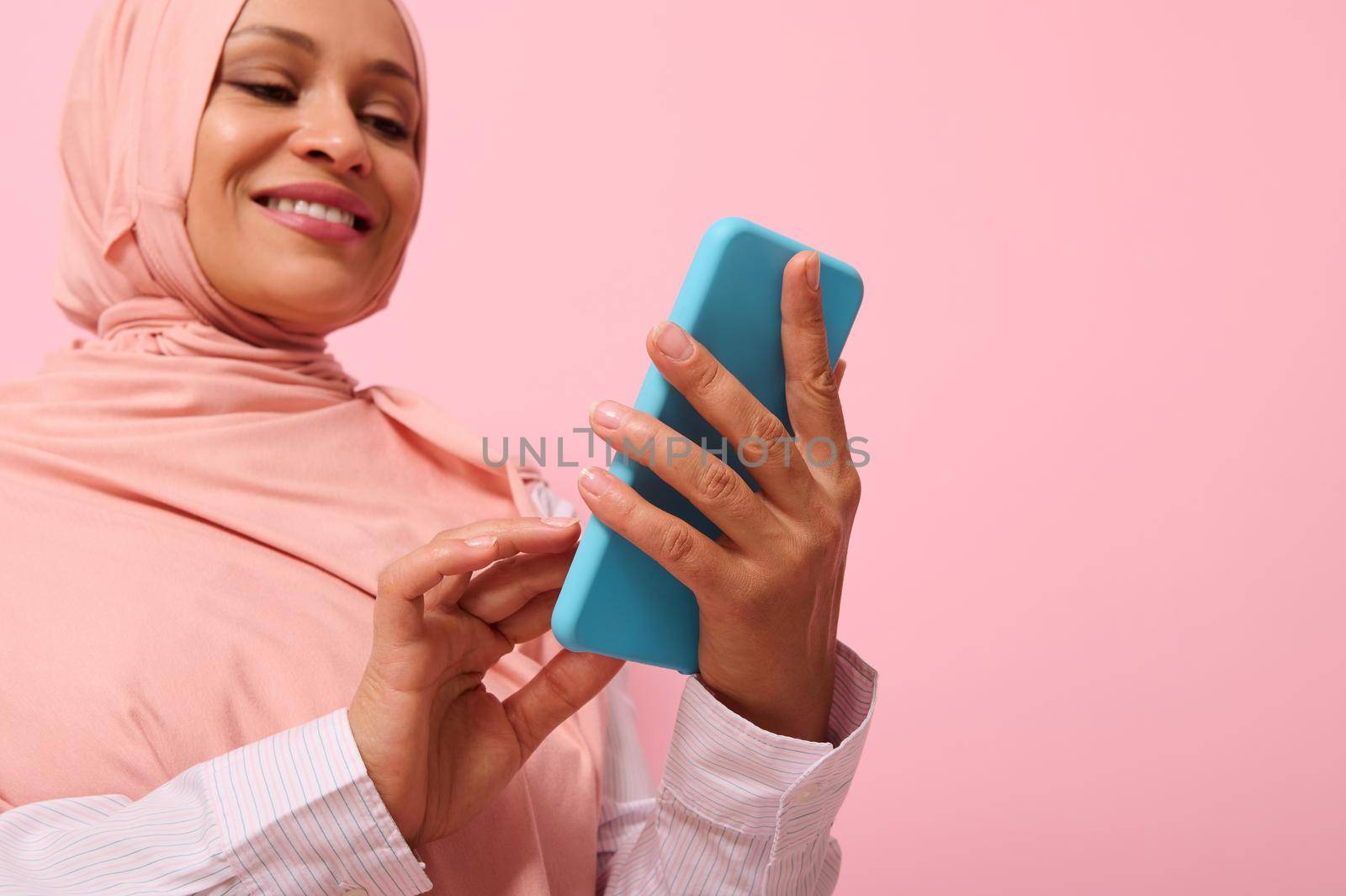 Smiling with toothy smile mature Muslim woman of Middle Eastern ethnicity in pink hijab with smartphone in blue cover on her hands, isolated on pink pastel background with copy space for promotion