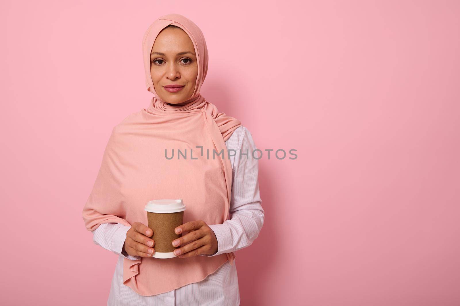 Arab Muslim beautiful woman wearing pink hijab and white shirt holding a recyclable disposable ecological paper mug in her hands, looking at camera posing on colored background with copy space