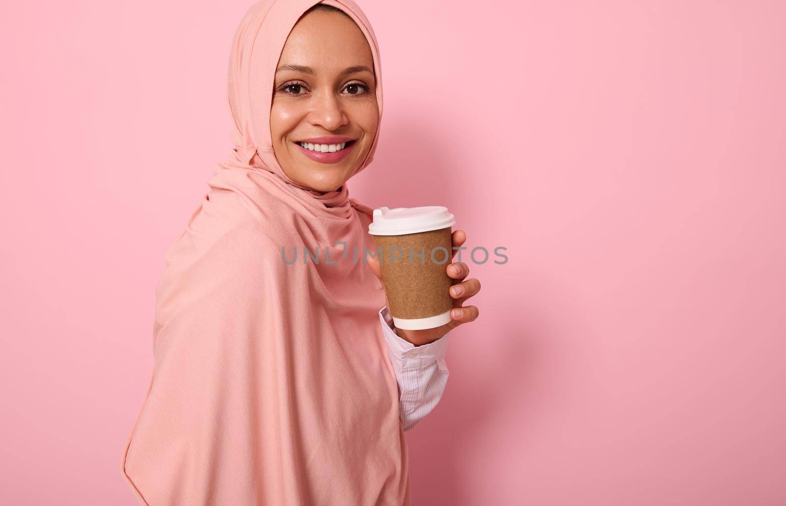 Close-up. Arabic Muslim woman with covered head in hijab holds disposable cardboard takeaway cup, smiles toothy smile, looking at camera, standing three quarters against colored background, copy space by artgf