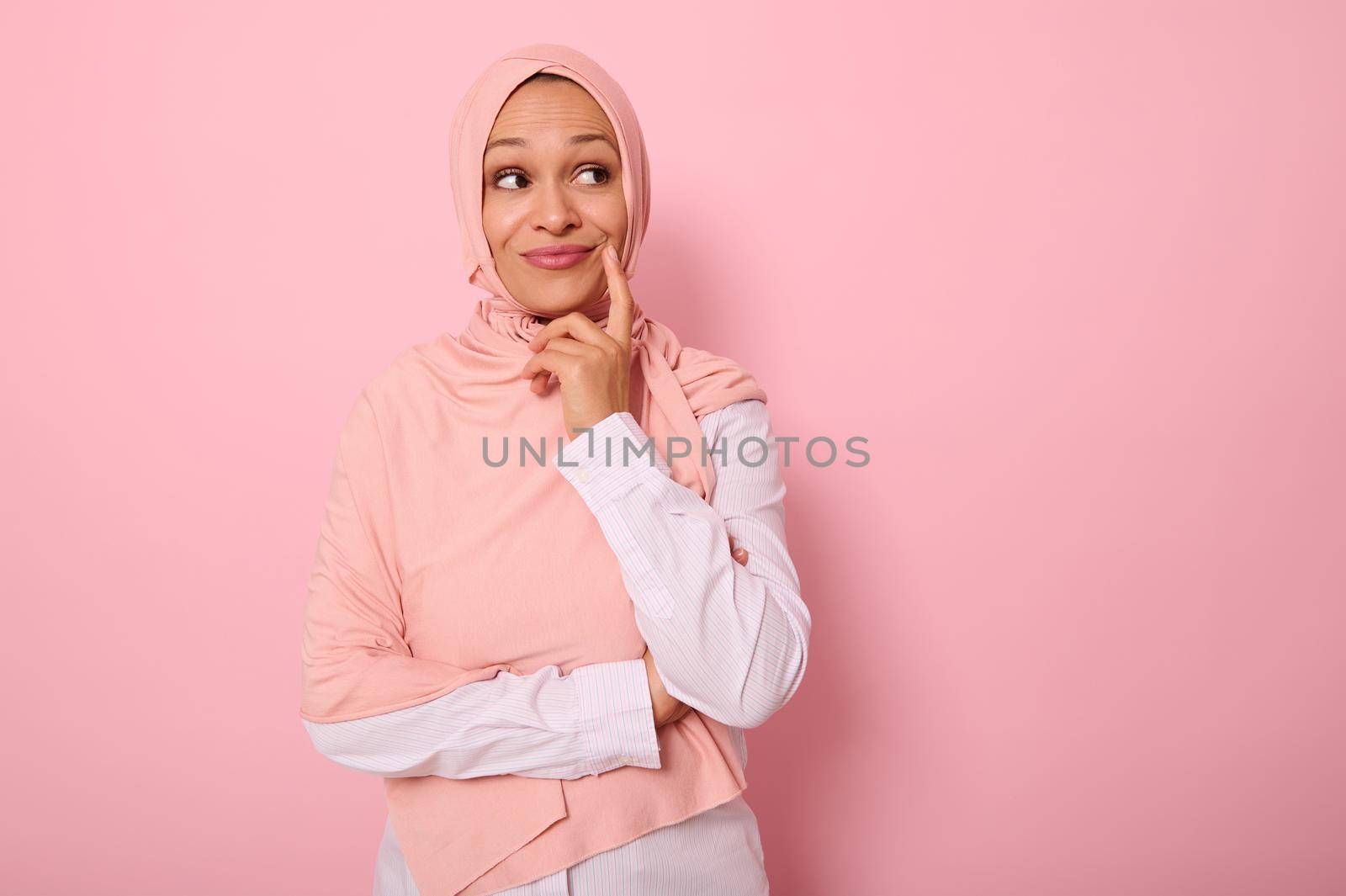 Adorable young Muslim beautiful gorgeous woman with a covered head in hijab looks mysteriously at a pink background with copy space and puts her finger to the corner of her lips, smiling thoughtfully
