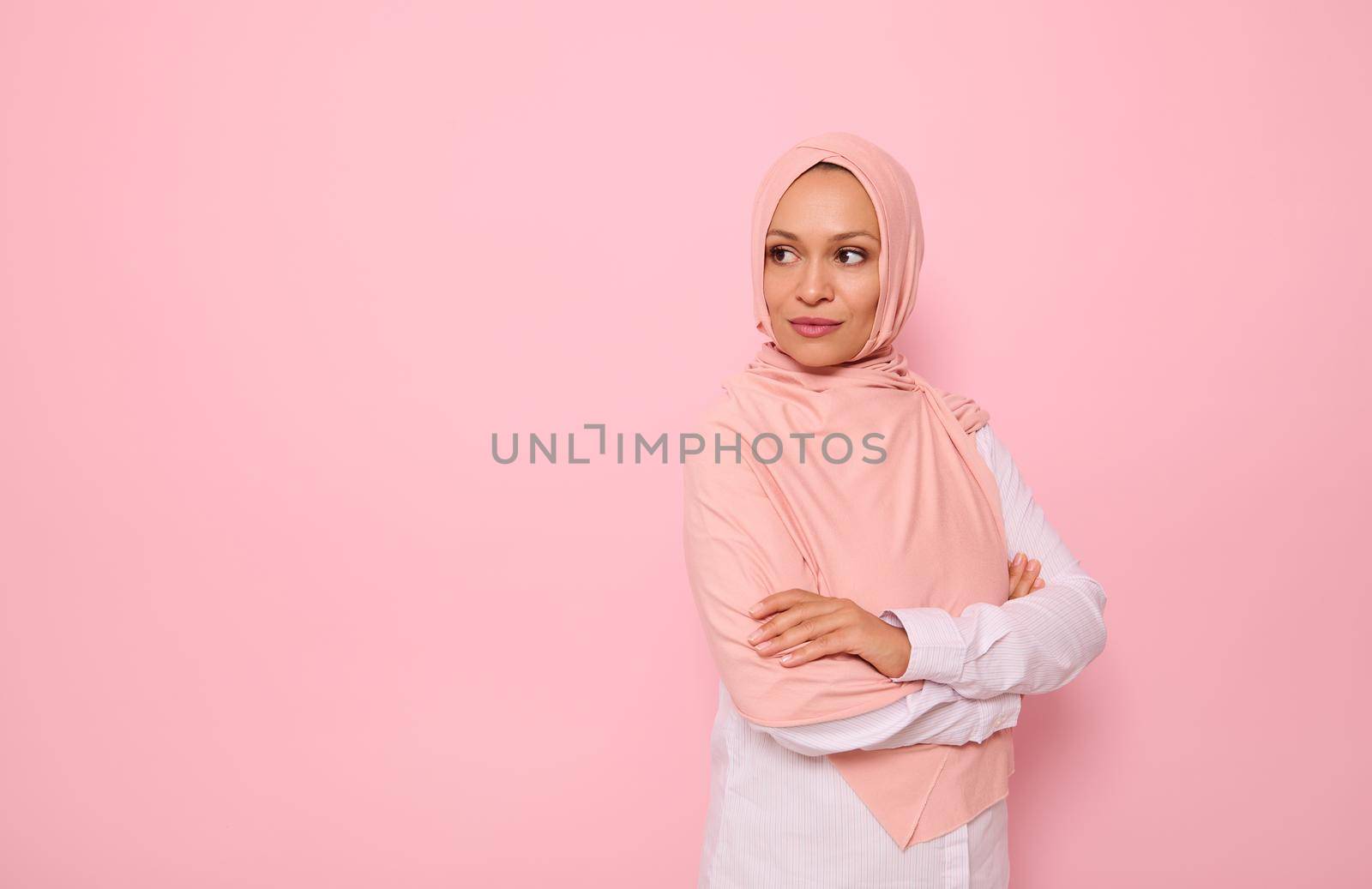 Confident portrait of Arabic Muslim gorgeous woman with attractive gaze and covered head with pink hijab, stands three quarters to colored background with copy space and looks away at copy space