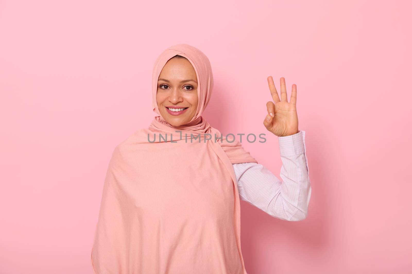 Charming joyful young Arab Muslim pretty woman dressed traditional religious attire and covered head in hijab showing OK sign, looking at the camera, standing against pink background with copy space