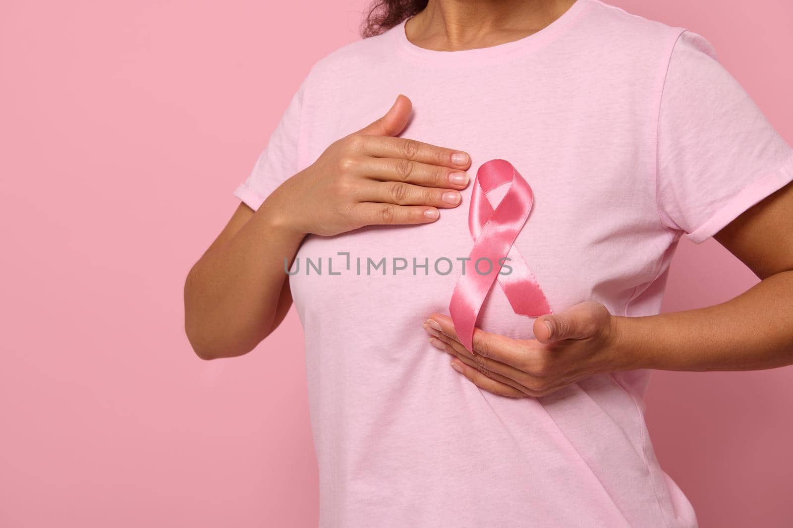 Close-up woman puts hands under pink ribbon on her T Shirt, supporting Breast Cancer Awareness campaign. Concept of 1 st October Pink Month, women's health care, colorecd background, copy space