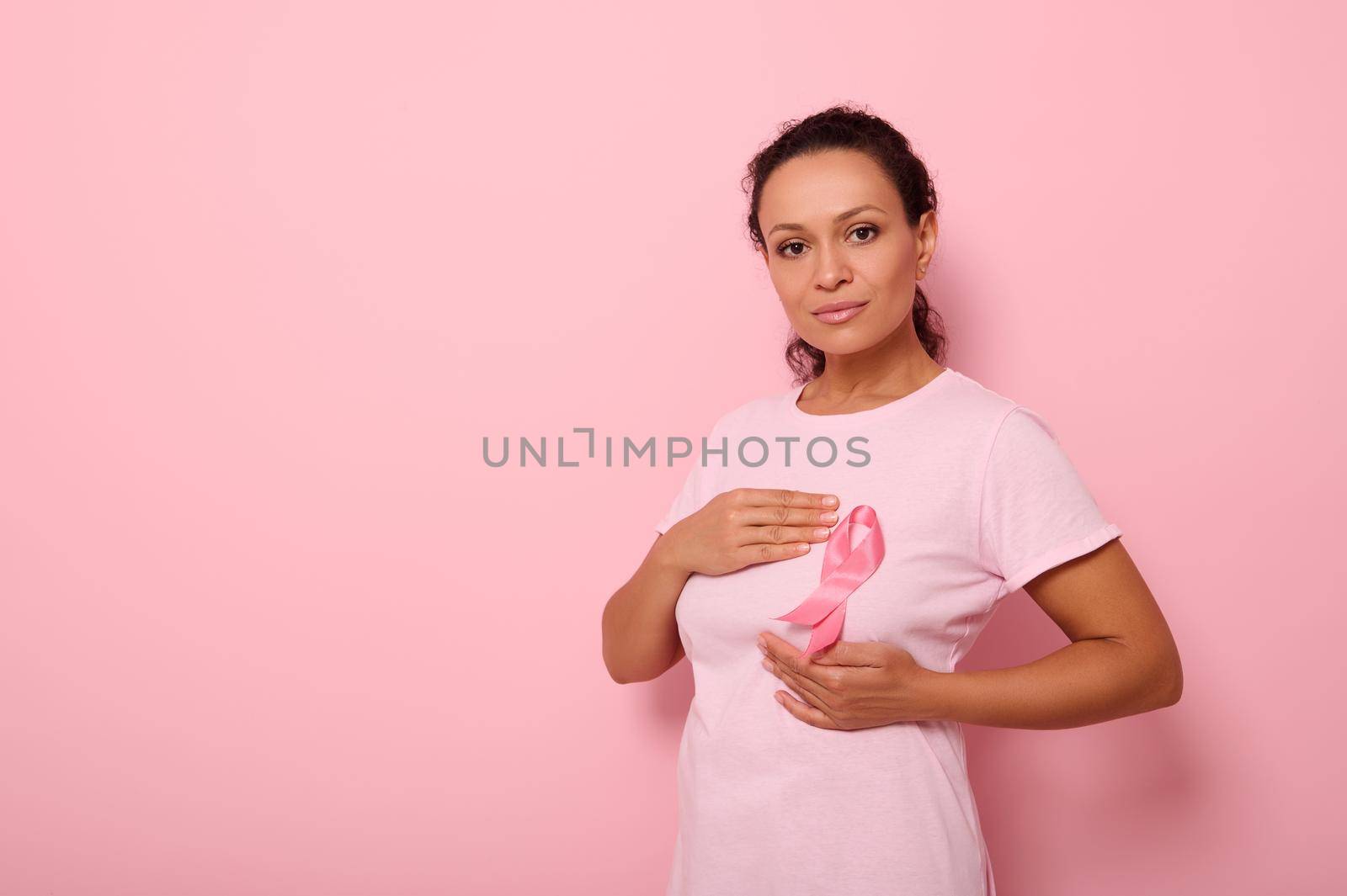 African American woman puts hands around pink ribbon on her pink T Shirt, for breast cancer campaign, supporting Breast Cancer Awareness. Concept of 1 st October Pink Month and women's health care by artgf