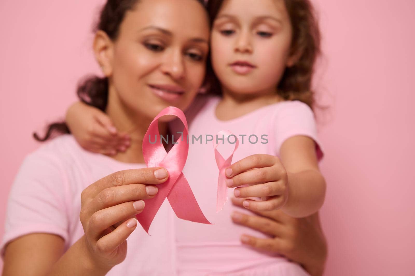 Close-up of pink satin ribbons on blurred woman and girl hugging each other. World Breast Cancer awareness Day, 1 st October. Women's health, medical concept, World Cancer Day, Cancer Survivor Day. by artgf
