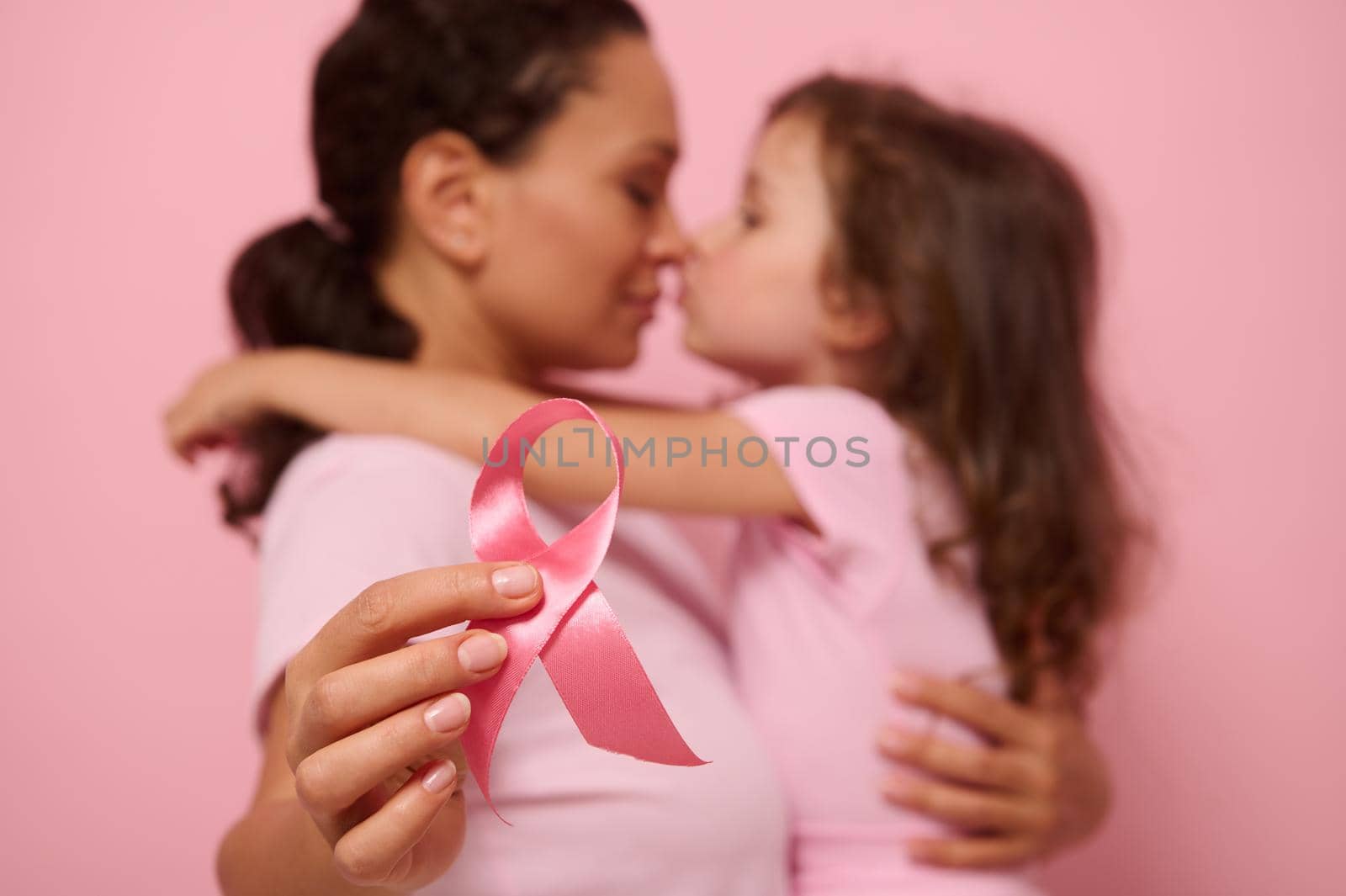 Soft focus on pink satin ribbon, symbol of International Breast Cancer Awareness Day, against blurred background of loving mother and lovely daughter. Female healthcare and medical education concept by artgf