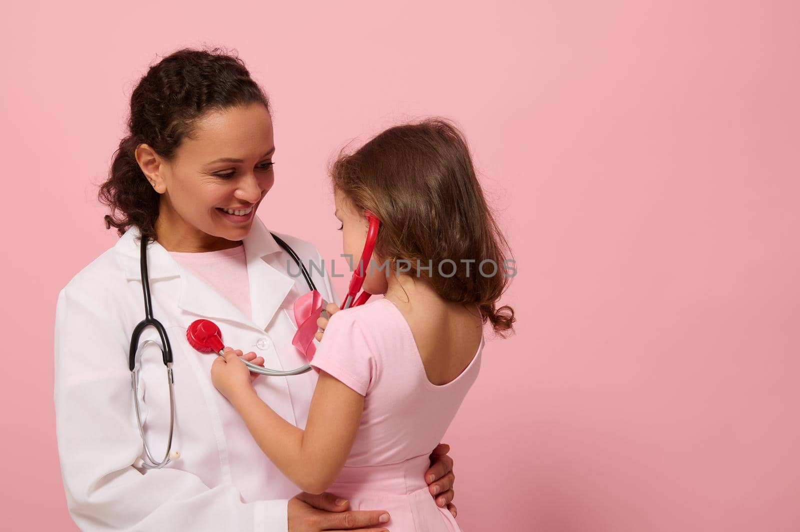 Little girl in pink clothes uses stethoscope, listens to chest and lungs breathing of doctor with pink ribbon on chest. Concept of educational program for supporting breast cancer patients. Copy space