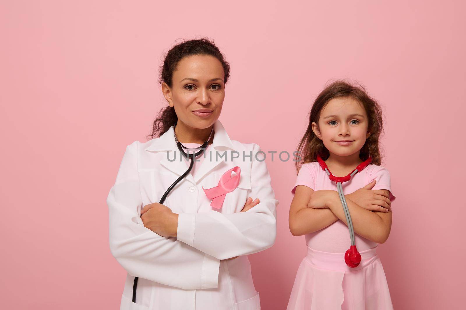 Gorgeous woman doctor in white medical gown stands next to a cute little girl in pink, both with crosses arms on chest and wearing a pink ribbon, symbol of Breast Cancer Awareness Day. Medical concept