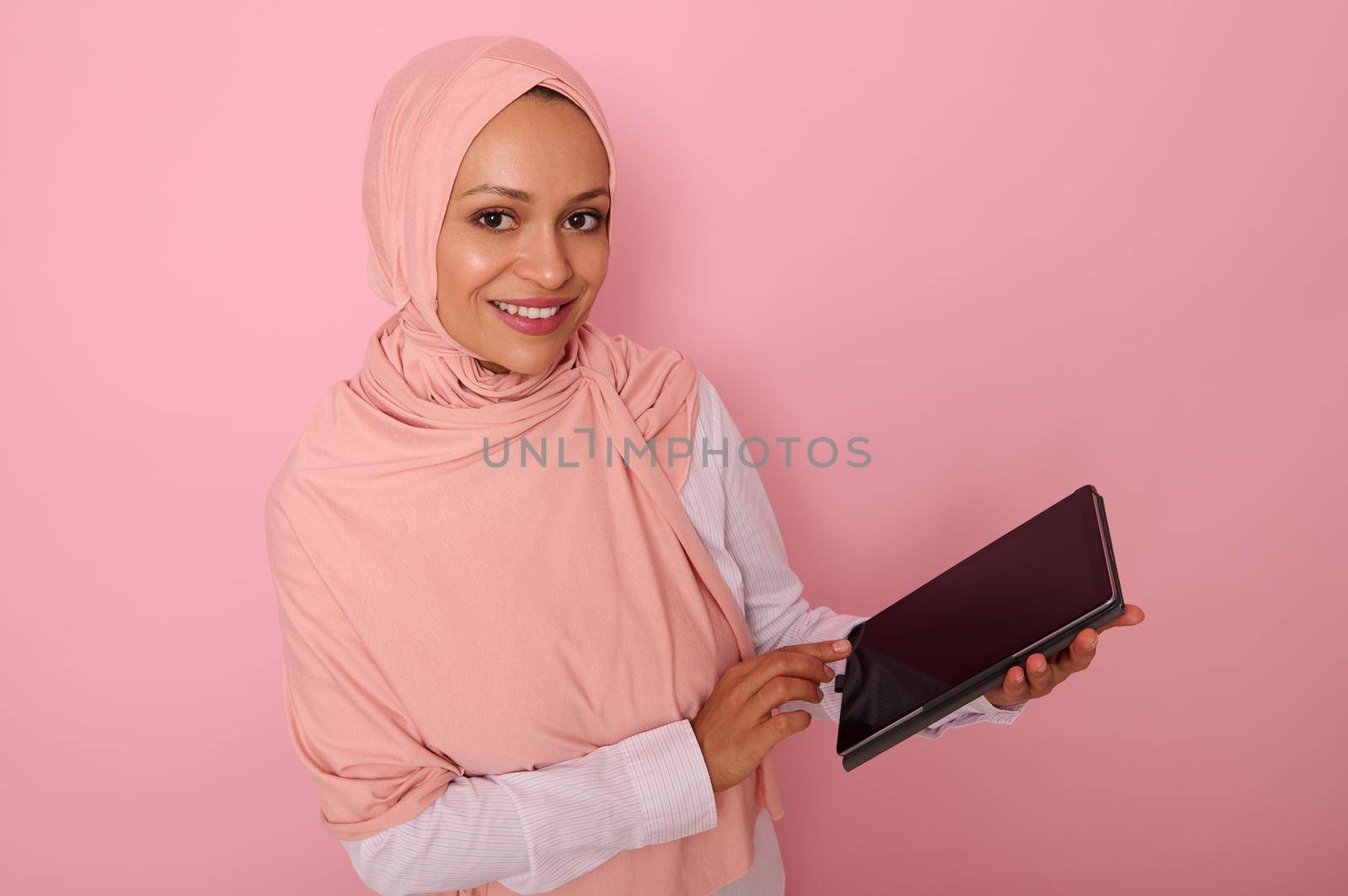 Arab Muslim woman in pink hijab smiles with toothy smile looking at camera, posing on colored pastel background with digital tablet in hands. Isolated portrait for advertising with copy space for text by artgf