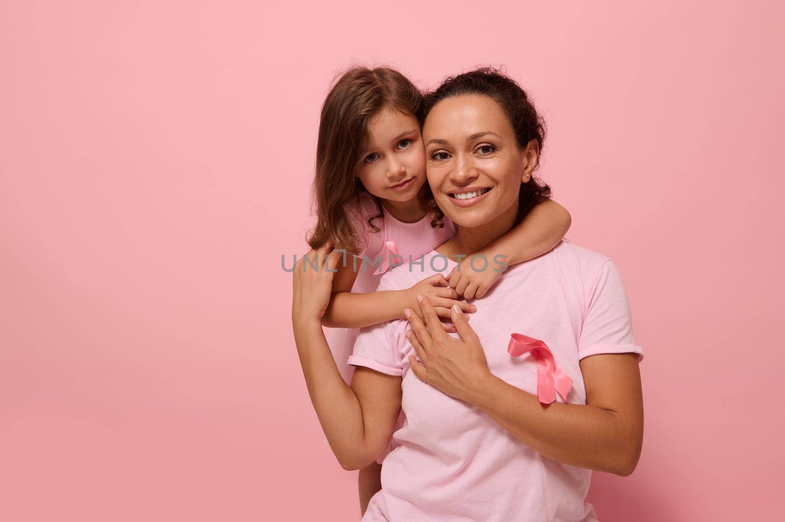 Woman's health , medical concept. Serene woman and girl in pink attire with Breast Cancer Awareness ribbon, daughter hugging her mom, holding by hands, looking at camera, supporting cancer patients. by artgf