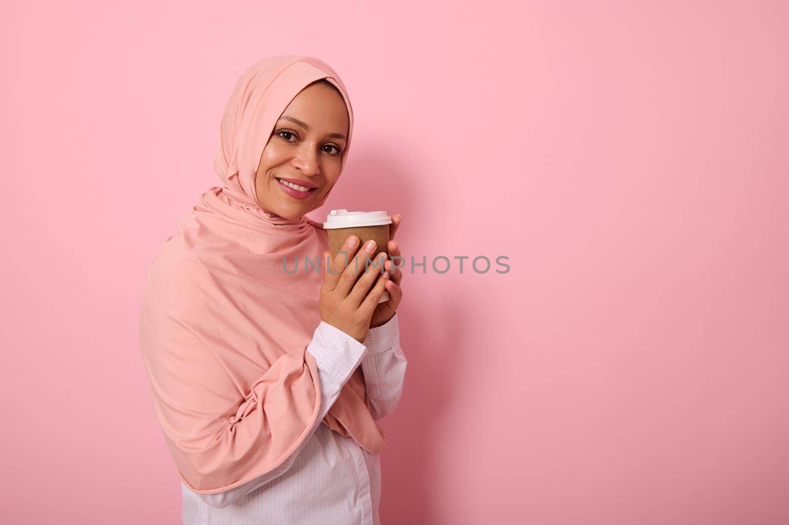 Arabic muslim pretty woman with covered head in pink hijab drinking hot drink, tea or coffee from disposable cardboard takeaway cup, standing three quarters against colored background with copy space by artgf