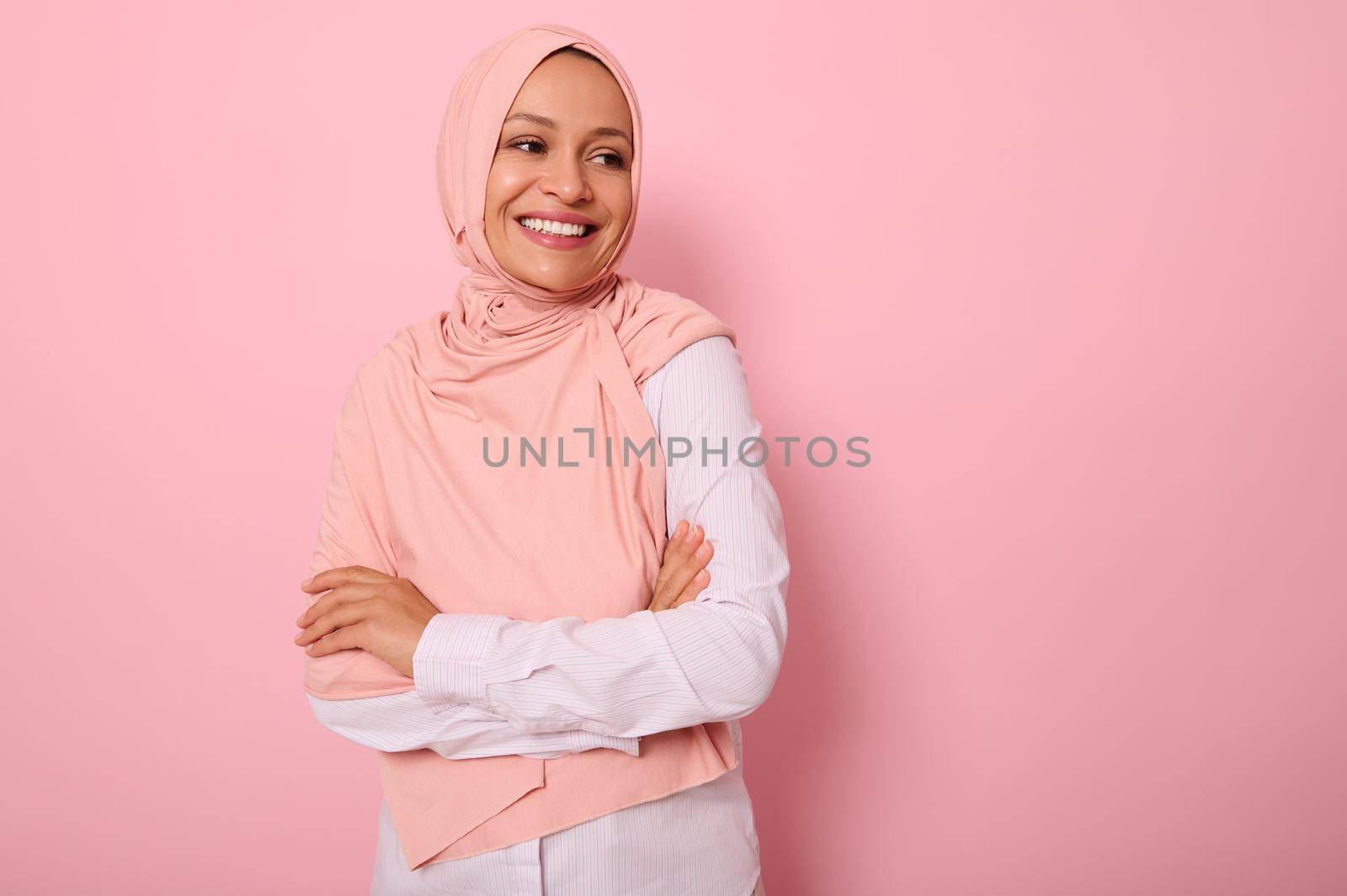 Waist-length portrait of beautiful Arabic Muslim woman in pink hijab posing looking to the side with attractive gaze, toothy smile, confident look and crossed arms, on pink background with copy space by artgf