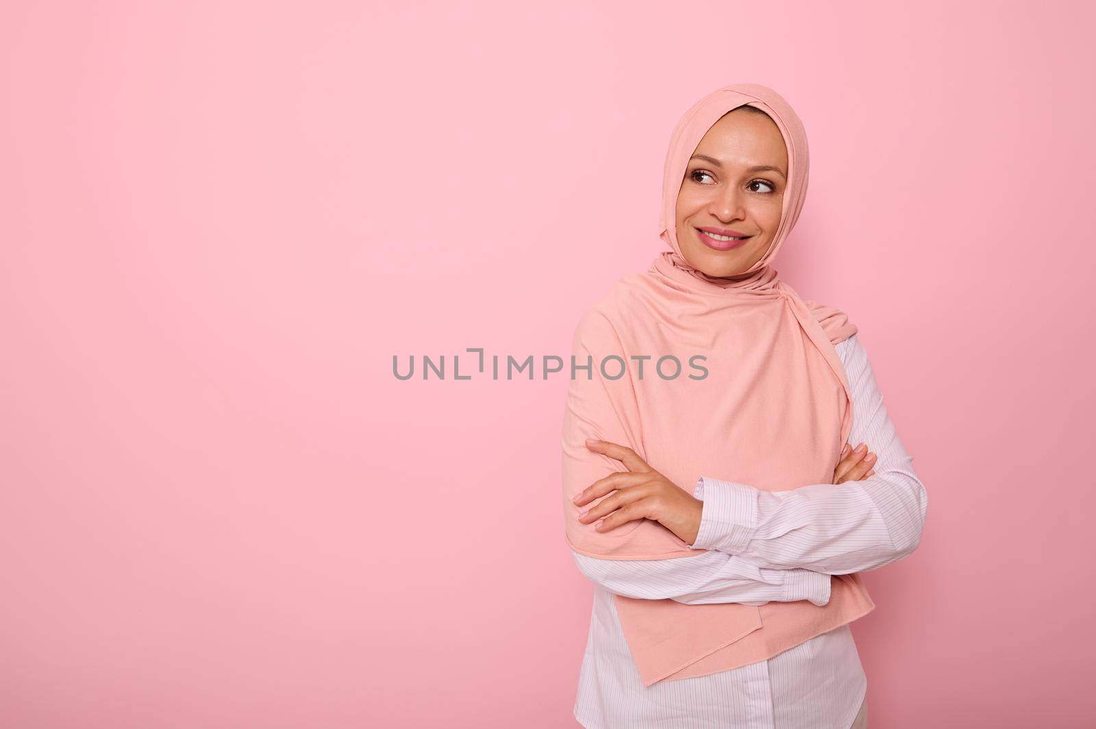 Waist-length portrait of an attractive young Arab Muslim gorgeous pretty woman with covered head in hijab looking mysteriously at the side on a pink background with copy space and smiling thoughtfully