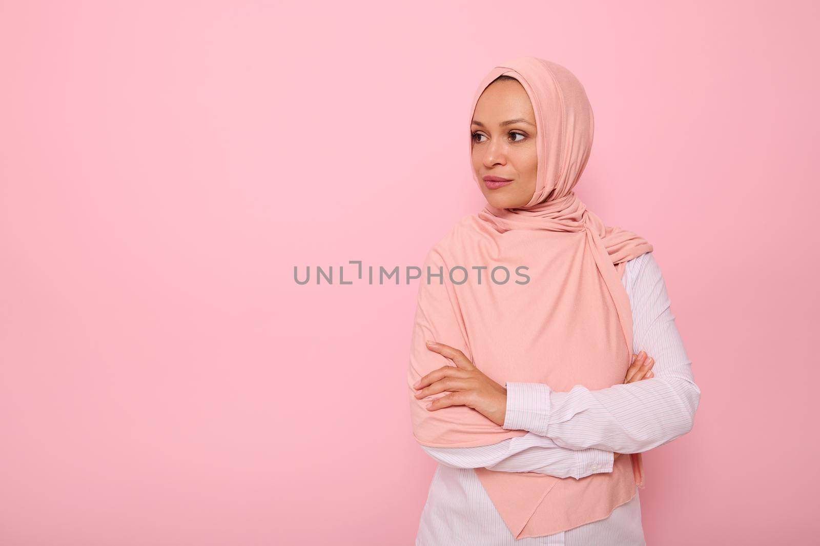 Confident portrait of Arabic Muslim beautiful woman with attractive gaze and covered head with pink hijab, stands three quarters to colored background with copy space and looks away at copy space