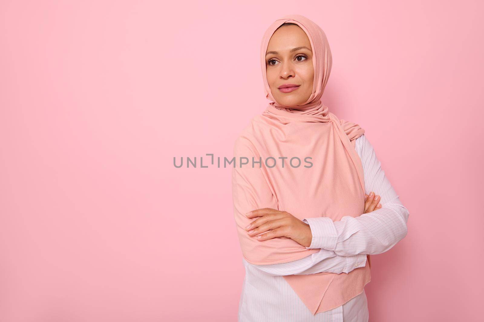 Confident portrait of Arabic Muslim beautiful woman with attractive gaze and look, wearing pink hijab and standing three quarters to colored background with copy space