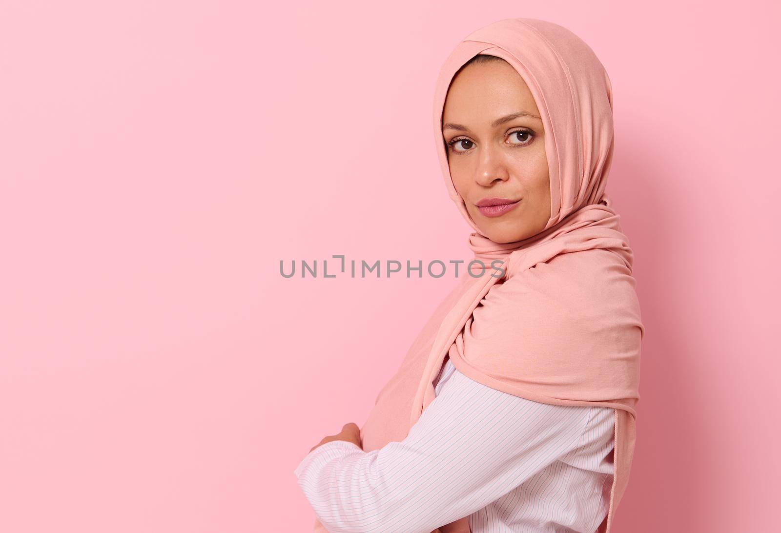 Confident close-up portrait of Arabic Muslim beautiful woman with attractive look and gaze, wearing a pink hijab, looking at camera confidently standing sideways against colored background, copy space