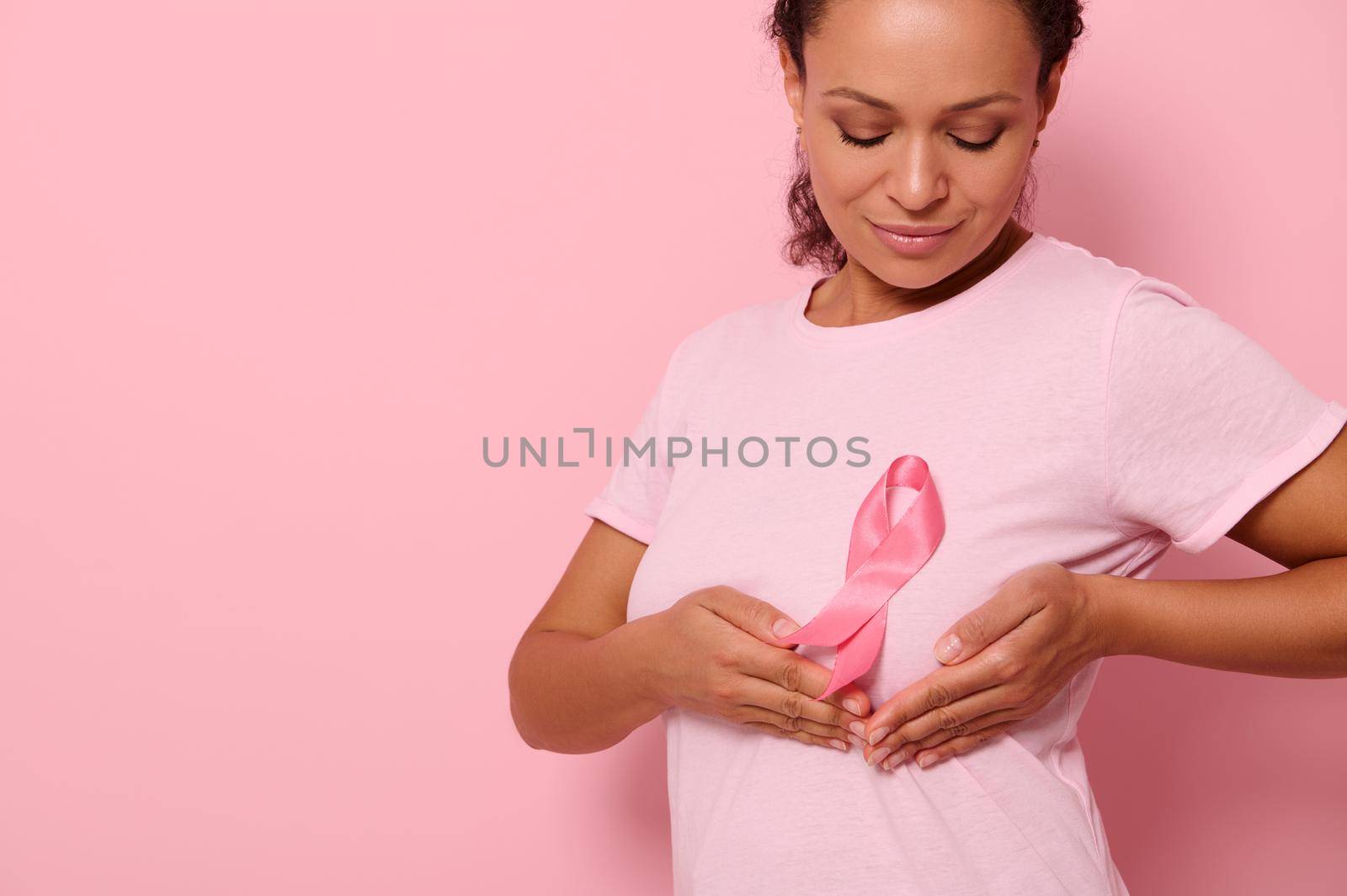 Close-up, mixed race woman puts hands under pink ribbon on her pink T Shirt, for breast cancer campaign, supporting Breast Cancer Awareness. Concept of 1 st October Pink Month and women's health care by artgf