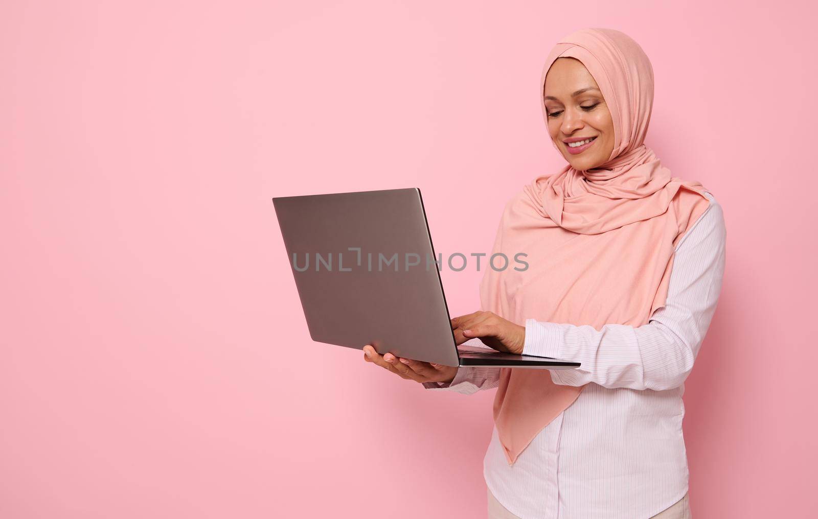 Portrait of a beautiful happy Muslim Arab woman in pink hijab working on laptop, isolated on colored background with copy space. Successful programmer, IT female worker, start-up, business lady