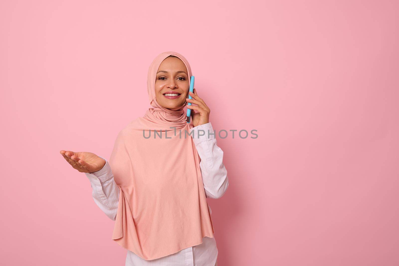 Muslim Arab gorgeous woman with covered head in pink hijab talks on mobile phone and gestures with hands showing her palm to a copy space . Isolated portrait on pink pastel background for advertising