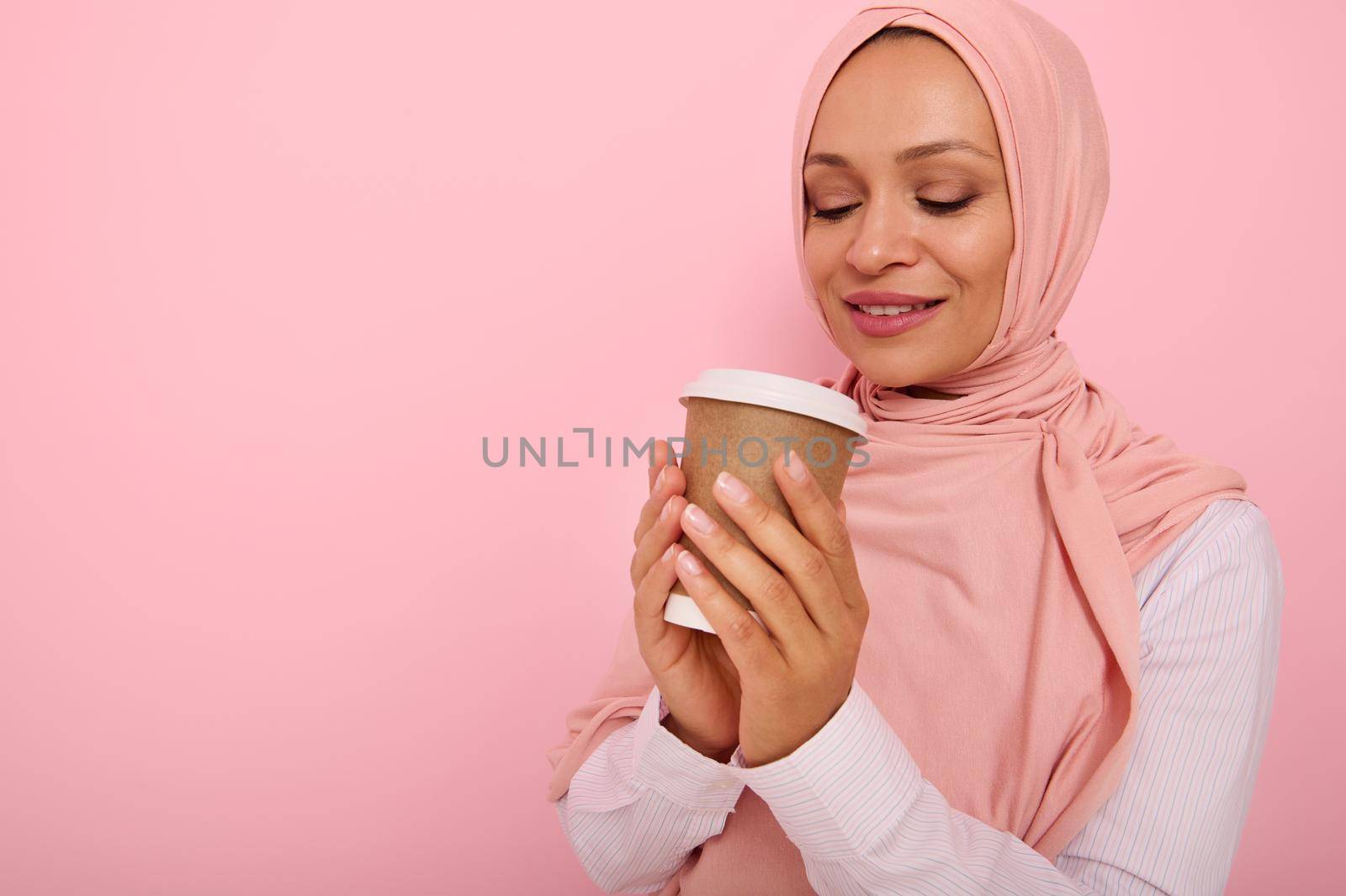 Beautiful Arabic muslim woman with covered head in pink hijab drinking hot drink, tea or coffee from disposable cardboard takeaway cup, standing three quarters against colored background, copy space by artgf