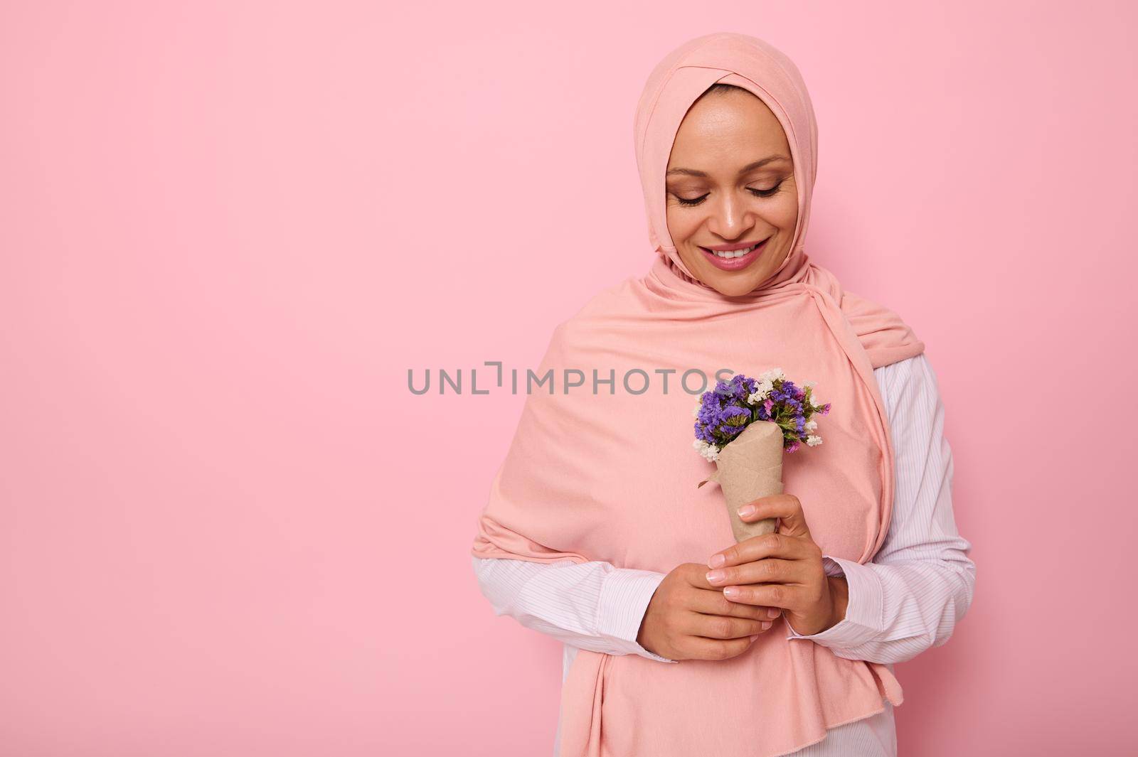 Isolated confident portrait on colored background with copy space of an attractive Muslim Arab woman in pink hijab, looking at a cute bouquet of wildflowers in purple shades, wrapped in craft paper by artgf