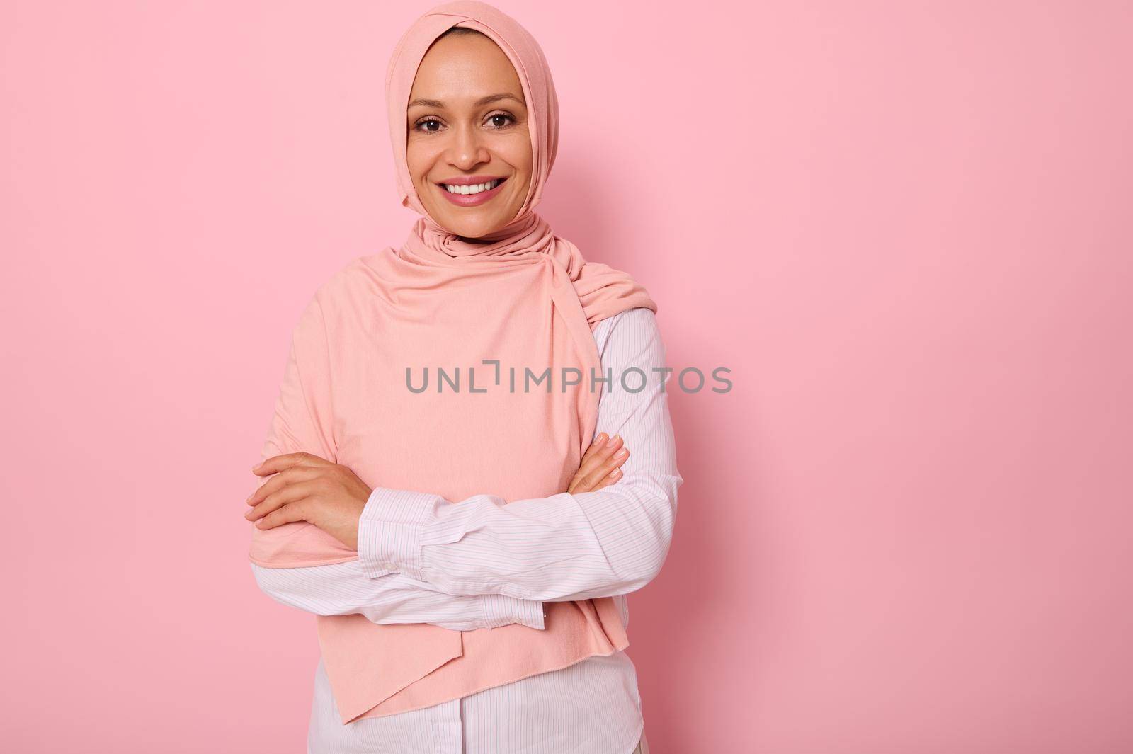Waist-length portrait of a beautiful Arabic Muslim woman in pink hijab posing looking to camera with attractive gaze, toothy smile, confident look and crossed arms, on pink background with copy space