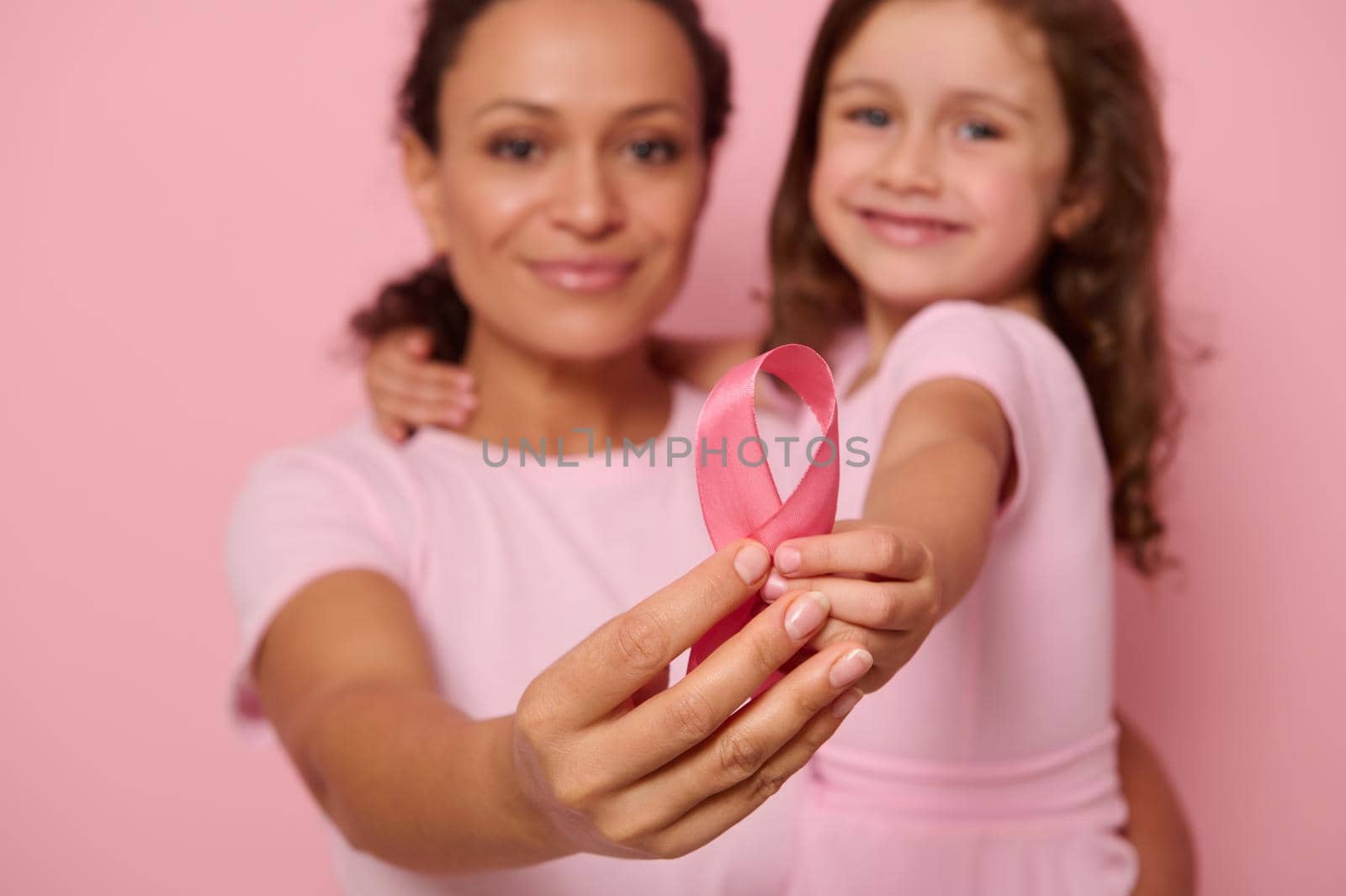 Focus on pink satin ribbon in the hands of two generations of women, mother and daughter posing on pink background, showing support, solidarity to patients cancer survivors. World Day of fight cancer by artgf